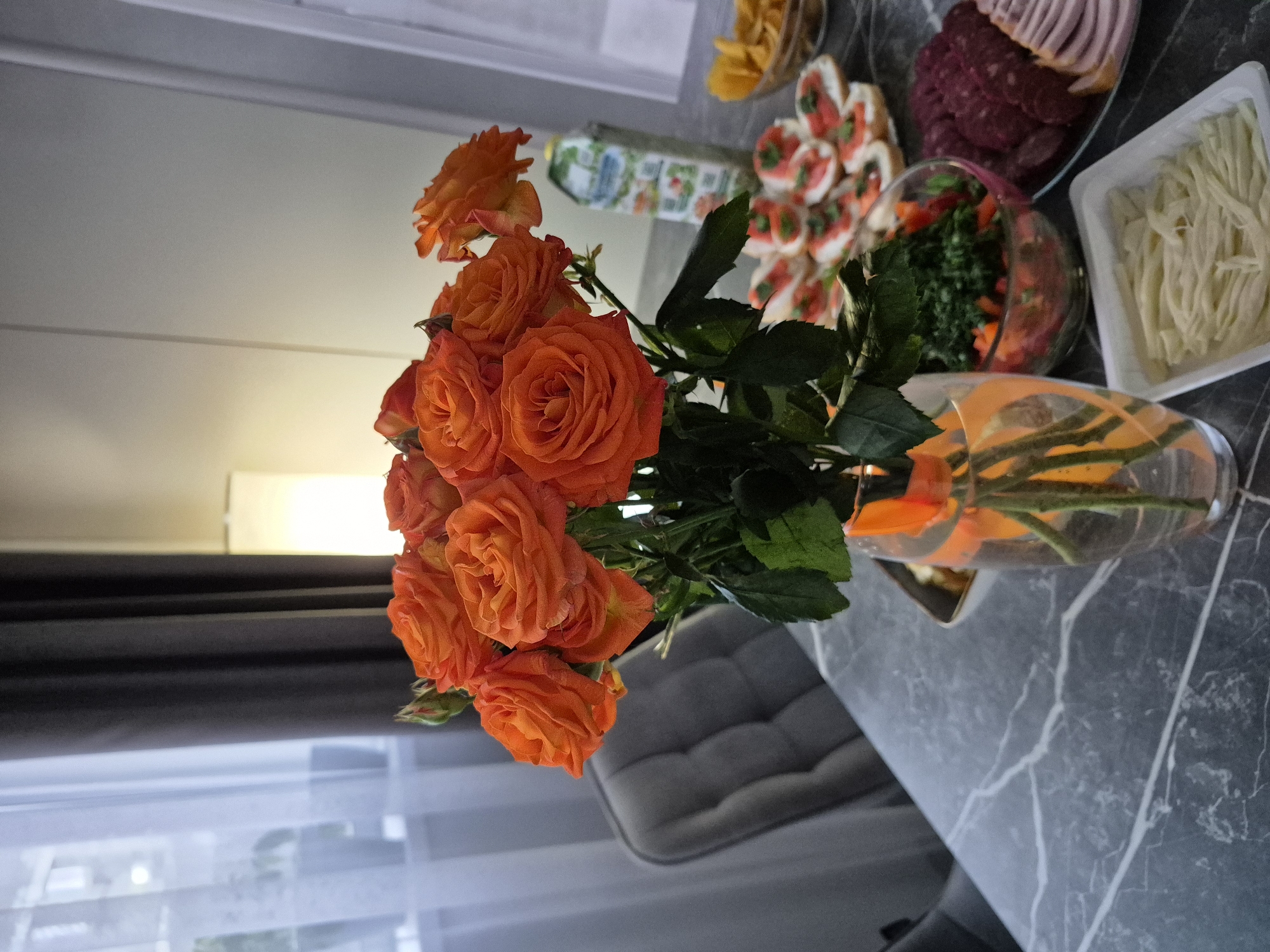 A table is set with various foods, including sliced meats, canapés, salad, and sliced cheese. An orange rose bouquet in a vase is featured prominently. The location is Chaikovsky, Russia. The table has a dark gray marbled surface. A cushioned chair is partially visible in the background.