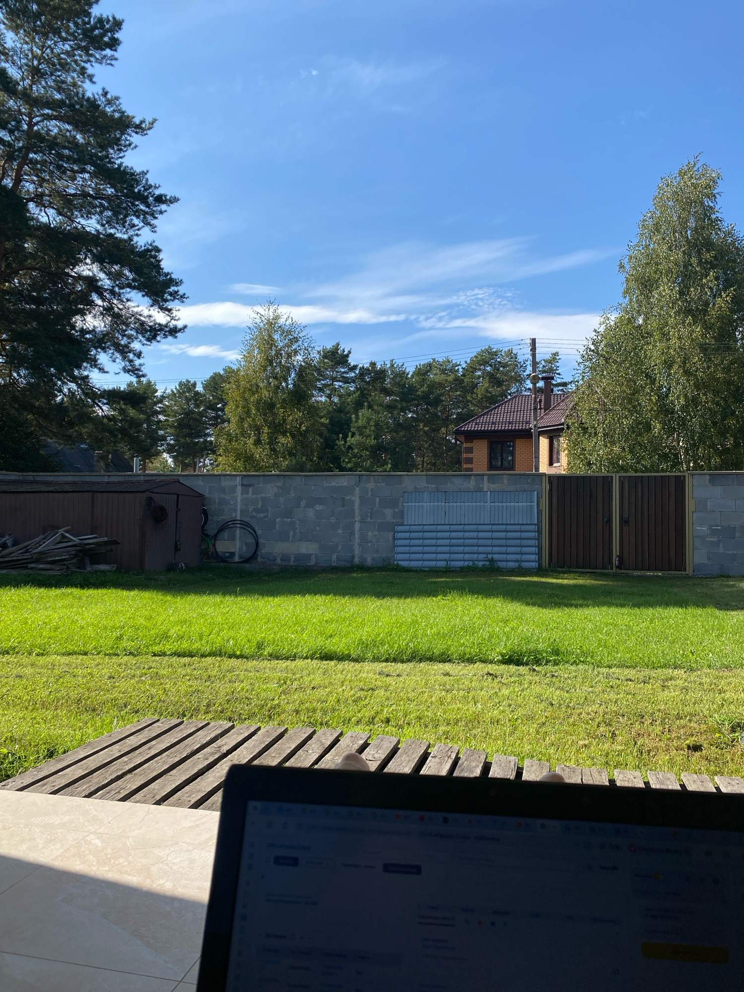 A sunny outdoor scene features a laptop in the foreground, its screen displaying text. Beyond the laptop, a wooden platform sits on a tiled surface, leading to a vibrant green lawn. In the middle ground, a grey concrete block wall runs horizontally across the frame. To the left of the wall, a dark brown shed stands next to a pile of lumber. A bicycle wheel is visible leaning against the wall. A section of the wall is covered with corrugated metal sheeting, and to its right, a dark brown wooden gate is installed. Behind the wall, a residential house with a brown tiled roof is visible, surrounded by lush green trees. The sky above is a clear blue with scattered white clouds. The location is specified as Perm, Russia.