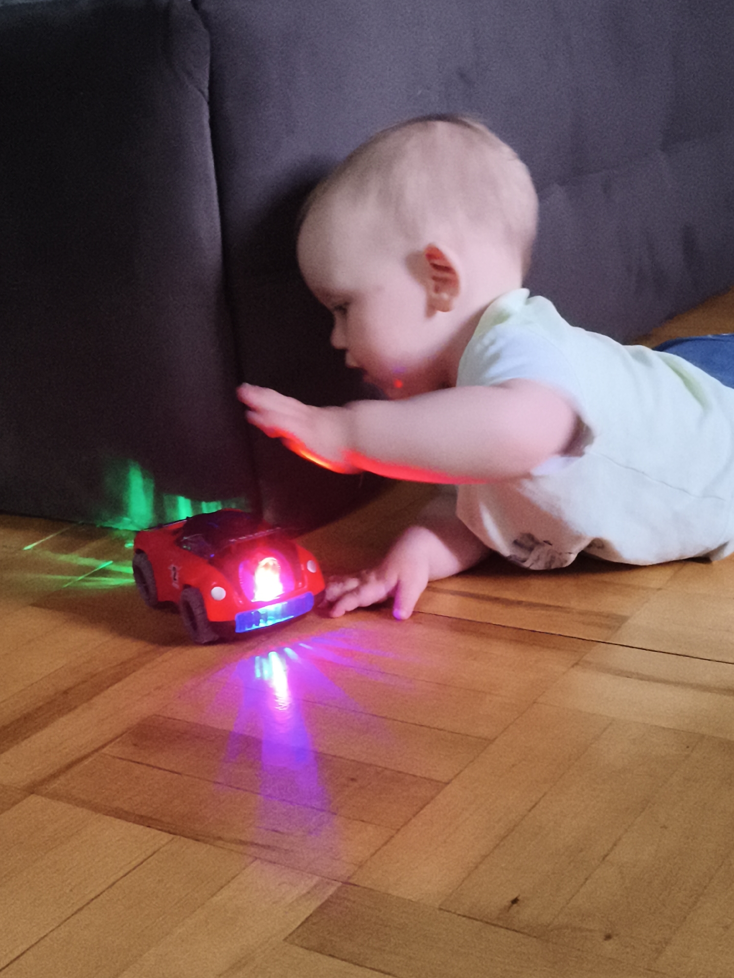 A baby is crawling on a wooden floor, reaching for a toy car. The car is red and has flashing lights in blue, pink, and green, casting colorful reflections on the floor and a dark surface, possibly a couch. The baby is wearing a light-colored t-shirt and jeans. The scene appears to be indoors, likely in a home, given the wooden floor and furniture. The lighting suggests it could be daytime, as the natural light from windows might be contributing to the scene's illumination, or it could be evening with artificial lighting. There is no visible text in the image. The baby's focused gaze and outstretched hand indicate engagement with the toy.