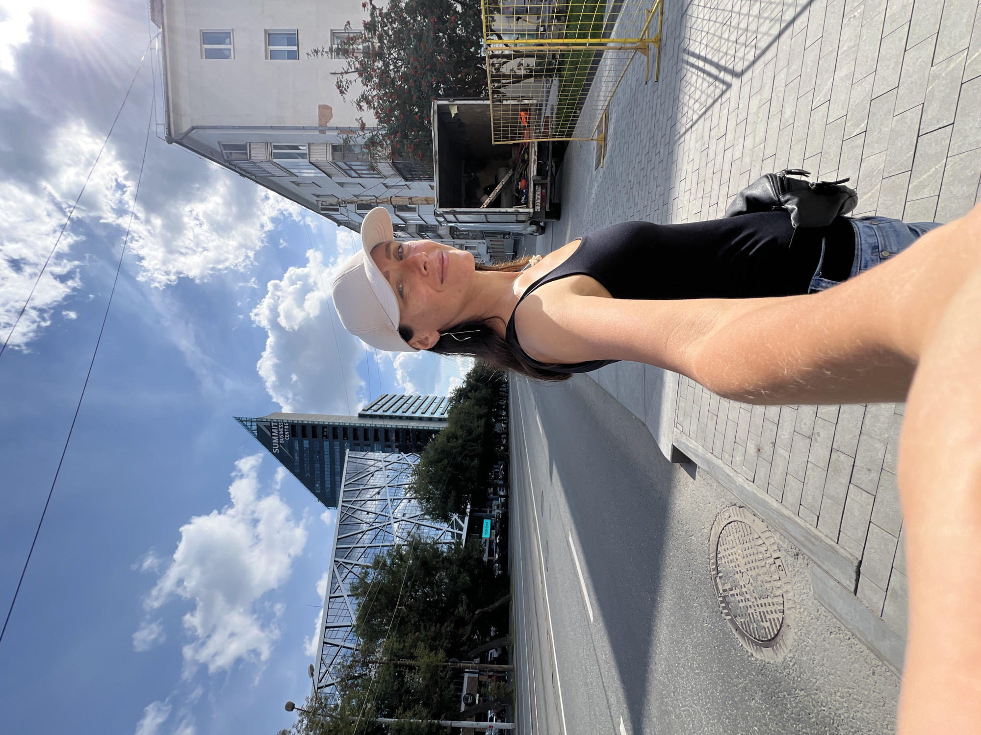 A woman wearing a white baseball cap and a black tank top takes a selfie on a sunny day in Yekaterinburg, Russia. She is standing on a sidewalk made of grey pavers next to a street. Behind her, a modern glass building with a triangular peak, labeled "SUMMIT BUSINESS CENTRE," is visible. To the right, a multi-story apartment building with balconies stands next to a white truck with an open back, suggesting a delivery or moving operation. Yellow safety fencing is also present on the sidewalk. The sky is blue with scattered white clouds.