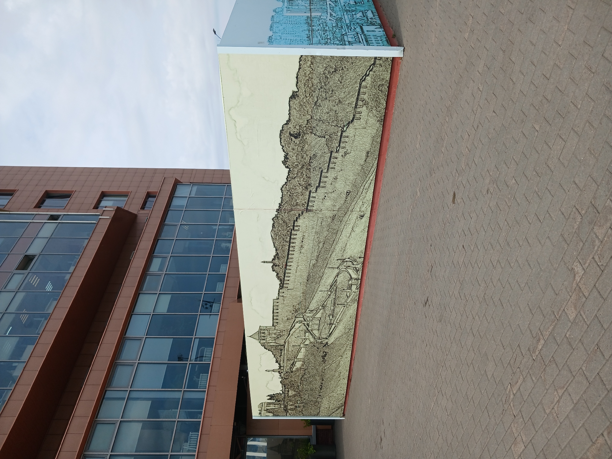 The image shows a large, rectangular mural or hoarding erected in front of a modern, multi-story building with brown cladding and large glass windows. The mural depicts a detailed, monochromatic sketch of the Nizhny Novgorod Kremlin, a historic fortress in the city of Nizhny Novgorod, Russia. The Kremlin is shown with its characteristic walls, towers, and surrounding landscape, rendered in a fine, intricate line drawing style with textured shading. The mural wraps around a corner, and on the side, a lighter blue sketch of a modern cityscape is visible. The foreground is paved with gray interlocking bricks. The sky is overcast. No people are visible in the scene. The building appears to be a commercial or office building.