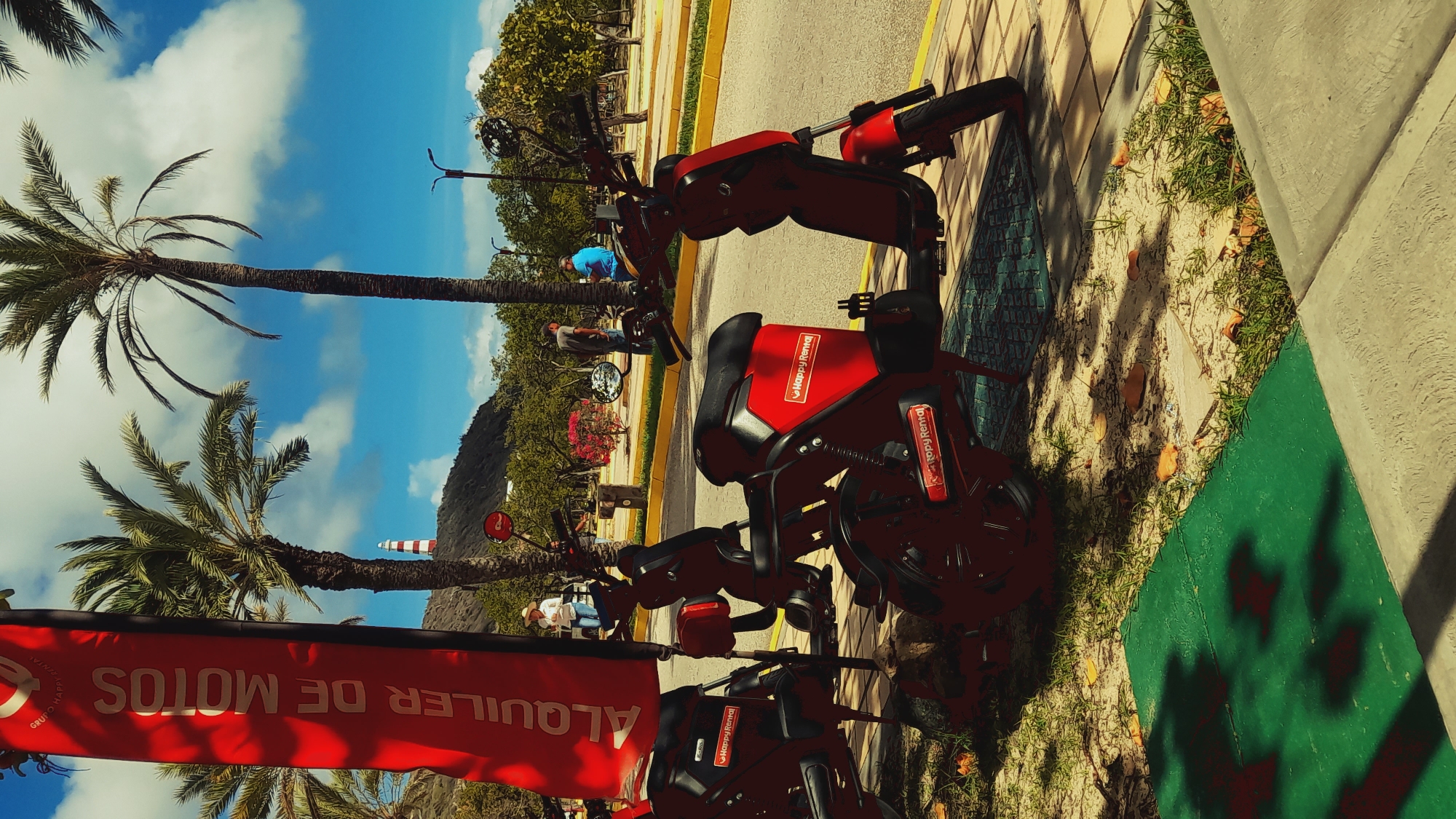 The image shows a row of red rental scooters parked on a sidewalk under palm trees, with a red banner on the left that reads "ALQUILER DE MOTOS" (Motorcycle Rental). In the background, there are people walking and standing, a lighthouse on a hill, and lush greenery. The scene suggests a sunny day in a warm, coastal location. The scooters have "Happy Rental" branding on them. The environment appears to be a public area with a paved road and a sidewalk. The location is specified as Pedro González, Venezuela.
