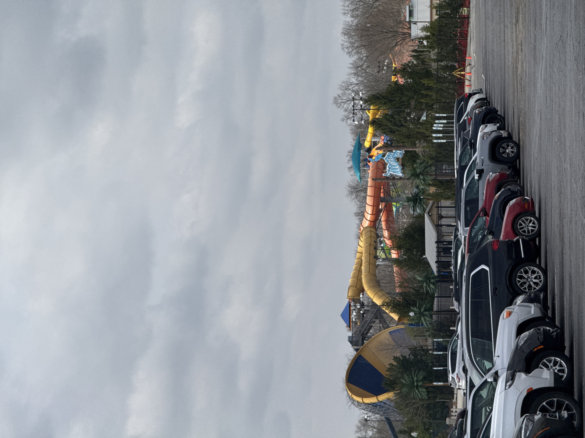 The image shows a parking lot filled with cars, with a water park visible in the background. The water park, identifiable as Zoombezi Bay based on the distinctive water slides, is located in Powell, Ohio, United States of America. The weather appears overcast. There are no people visible in the image. The prominent features are the water slides of the park and the cars in the parking lot. There are no interactions or actions taking place in the photo.