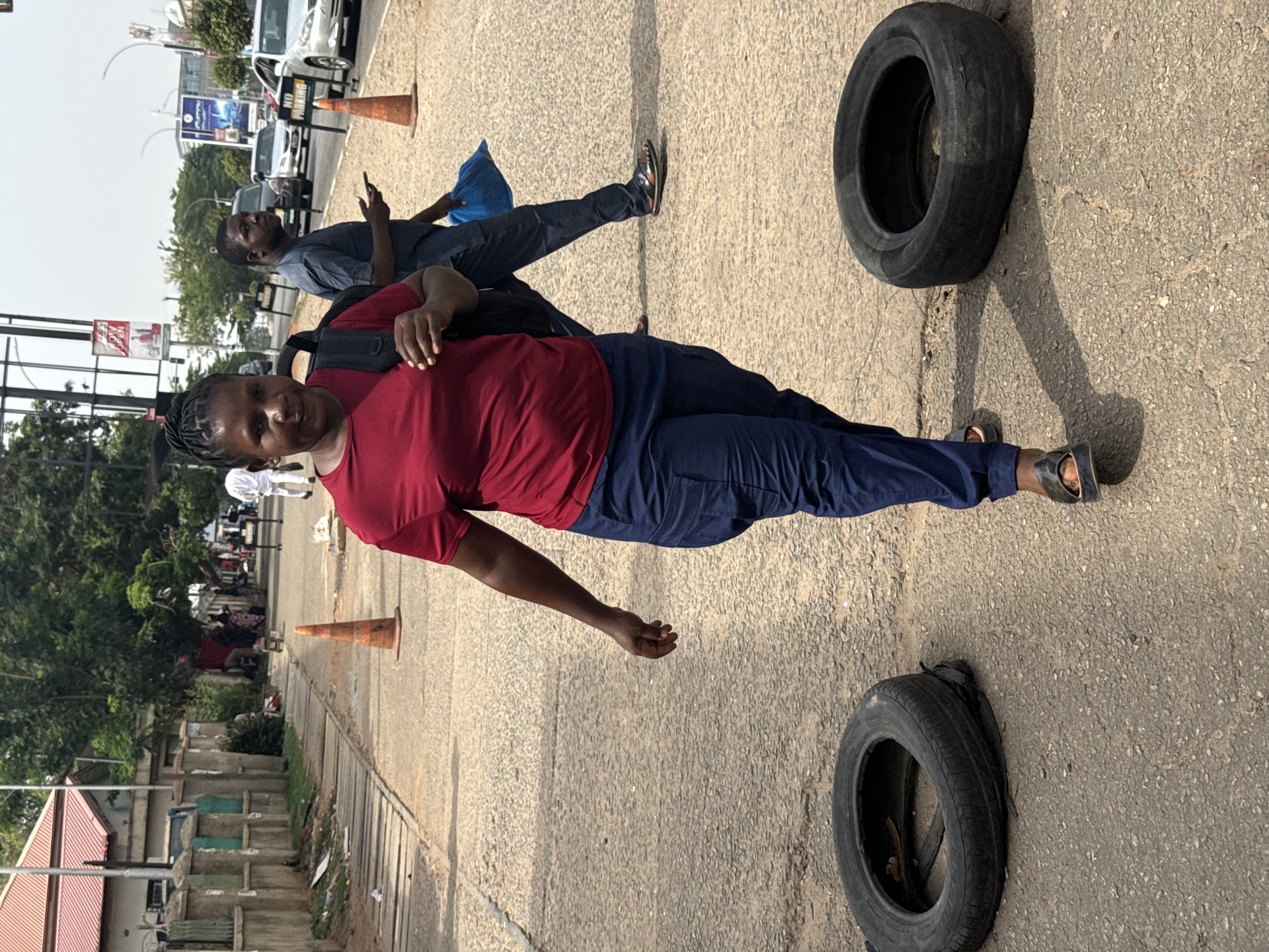 This image captures a bright, sunny daytime scene on a concrete or asphalt surface, likely a roadside or parking area. The strong sunlight casts clear, distinct shadows on the ground.
The primary subject is a woman in the foreground, positioned slightly off-center. She wears a short-sleeved red top, dark blue pants, and dark sandals. A black backpack is visible on her back. Her dark, braided hair is styled neatly. She is looking directly towards the camera with a subtle smile, and her right hand is slightly raised.
Behind and to her left stands a man, also wearing a dark blue t-shirt and dark pants. He appears to be looking upwards and slightly away from the camera, with one hand near his chest.
On the ground, two worn, black tires are visible; one in the upper right corner and another in the lower left. Two orange traffic cones are also present in the scene, one closer to the man and another further in the background.
The background reveals an urban or semi-urban environment with a street or road. Several vehicles, including cars and possibly a truck, are visible in the distance. Buildings with distinct red roofs and trees line the road. A white sign on a pole behind the man clearly displays the word "PARKING." Other billboards are present in the far background, but their text is illegible. The overall setting suggests a bustling area consistent with Abuja, Nigeria.