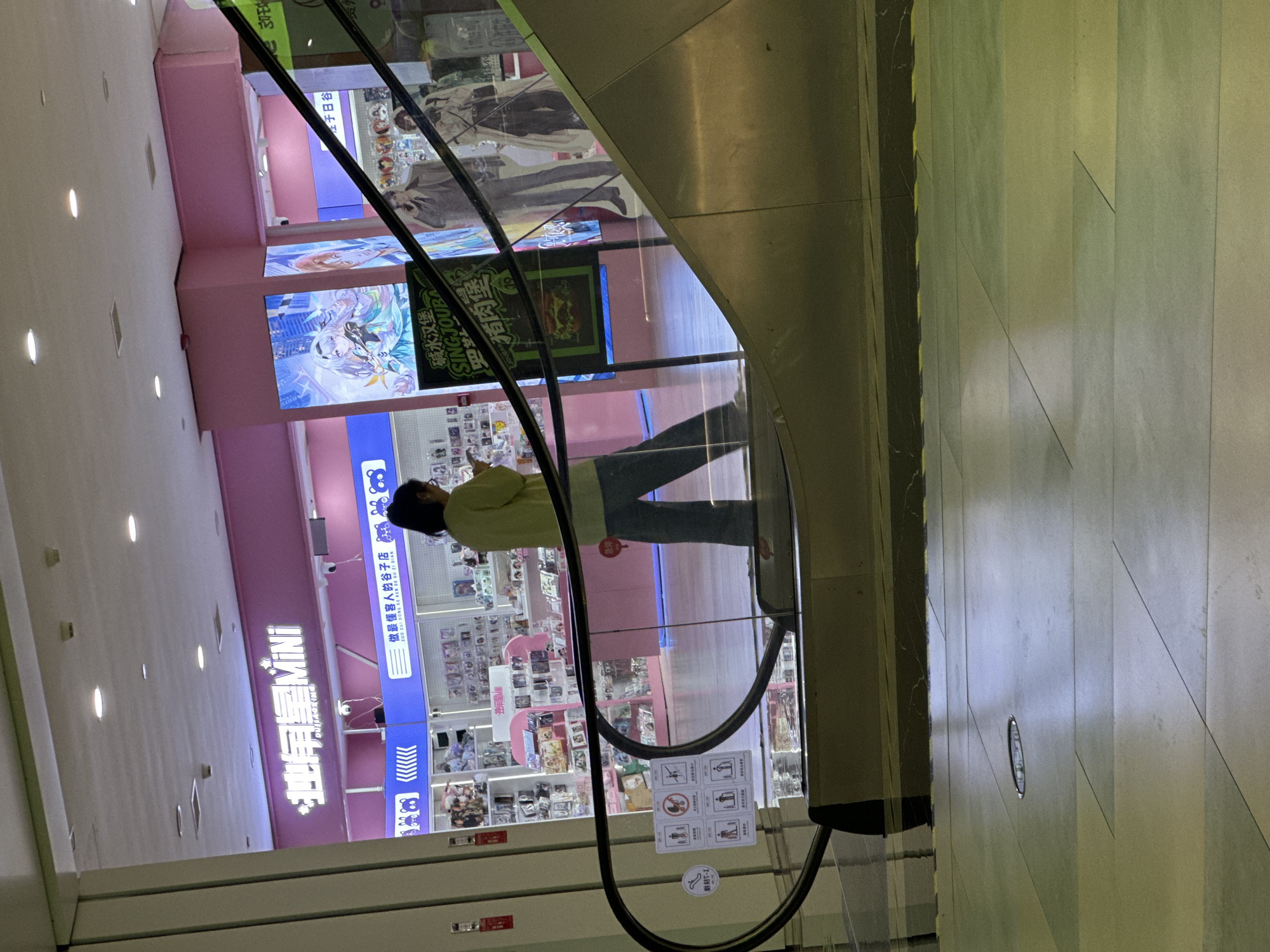 The image shows a shopping mall interior with an escalator and several shops. A person is walking on the escalator, looking at their phone. The shops appear to sell anime-related merchandise, with colorful displays and posters. The text visible on the signs suggests the location is in the Chongqing High-tech Zone, China. The lighting is a mix of overhead fluorescent lights and illuminated displays. The floor is tiled, and the escalator handrail is made of black rubber.