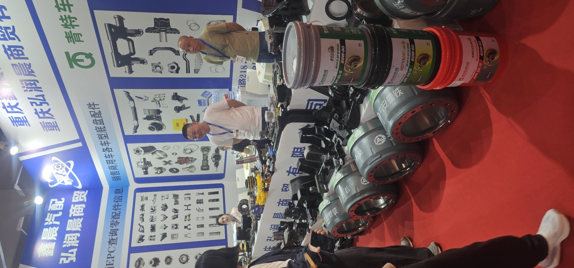 The image shows a trade show booth for a Chongqing-based company named “Xin Chen Auto Parts,” with the brand, "Hong Run Chen Trading." The booth features informational posters showcasing various auto parts and branding. Two men are visible standing behind the booth. The booth is displaying auto parts, including large metal casings and stacked buckets of oil labeled "Qingte Bridge Special Oil" and "API GL-5." The oil buckets have labels for 85W-90 and 80W-90. The backdrop of the posters and the red carpet beneath the products give the scene a business-like, professional atmosphere. The image might have been taken at a trade show event in the Liangjiang district of Chongqing, China, given the company's location.