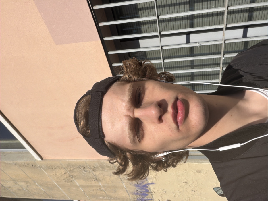 A young male with light brown, curly hair, wearing a black backward-facing baseball cap, is captured in a medium-close-up photograph. He wears a black jacket over a white t-shirt and white wired earphones. Two small silver hoop earrings are visible in his left ear. His eyes are squinted, and his lips are slightly parted, consistent with exposure to direct, bright sunlight. The background reveals an urban setting with two distinct building facades. To the left, a weathered yellow-beige wall exhibits texture and faded purple graffiti. To the right, a light peach-colored wall features a window secured by dark grey vertical metal bars, with horizontal blinds visible within the window recess. While the architectural style is consistent with urban areas of Milan, Italy, no specific landmarks are present to identify a precise location.