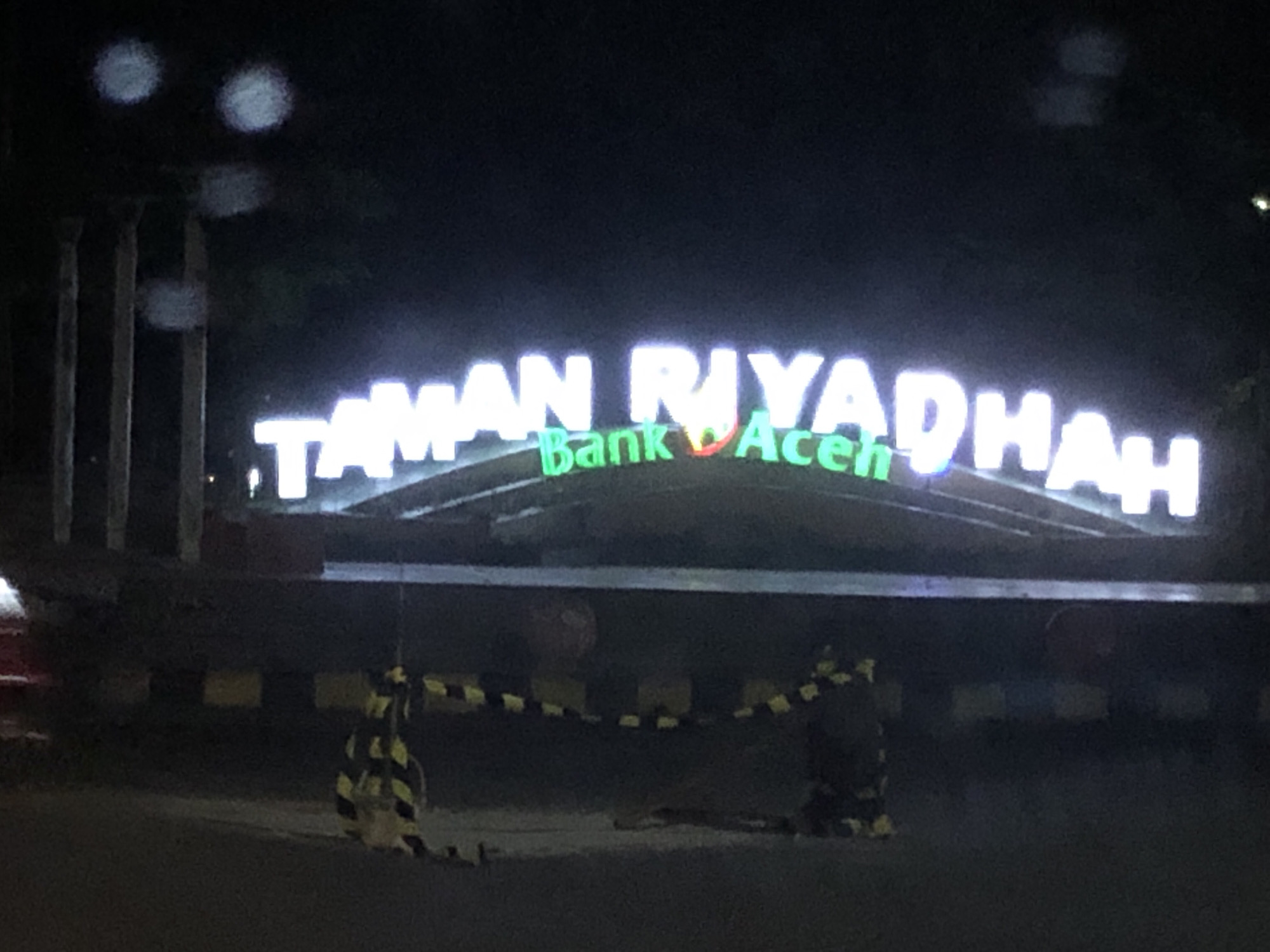 The image captures the entrance to Taman Riyadhah in Lhokseumawe, Indonesia, at night. The words "TAMAN RIYADHAH" are prominently displayed in illuminated white block letters, with "Bank Aceh" in green letters. A barrier, marked with black and yellow stripes, stretches across the foreground, likely controlling access. The background is dark, with blurry lights suggesting street lamps or vehicle headlights. Columns are partially visible on the left side. The scene appears still, without any people visible.