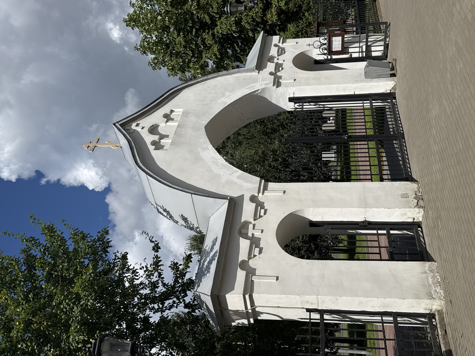 The image depicts an arched entranceway with black metal gates, likely belonging to a cemetery or religious institution, located in the city of Saint Petersburg, Russia. The entrance is constructed of white plaster and features architectural details such as decorative curves and a prominent cross atop the arched roof. Lush green trees frame the entrance, and a cloudy sky with patches of blue is visible. A black lamppost stands to the left, and a metal trash bin is situated to the right of the entrance. The ground is paved with light-colored bricks. The gates appear to be closed or partially open. There are no people visible in the image.