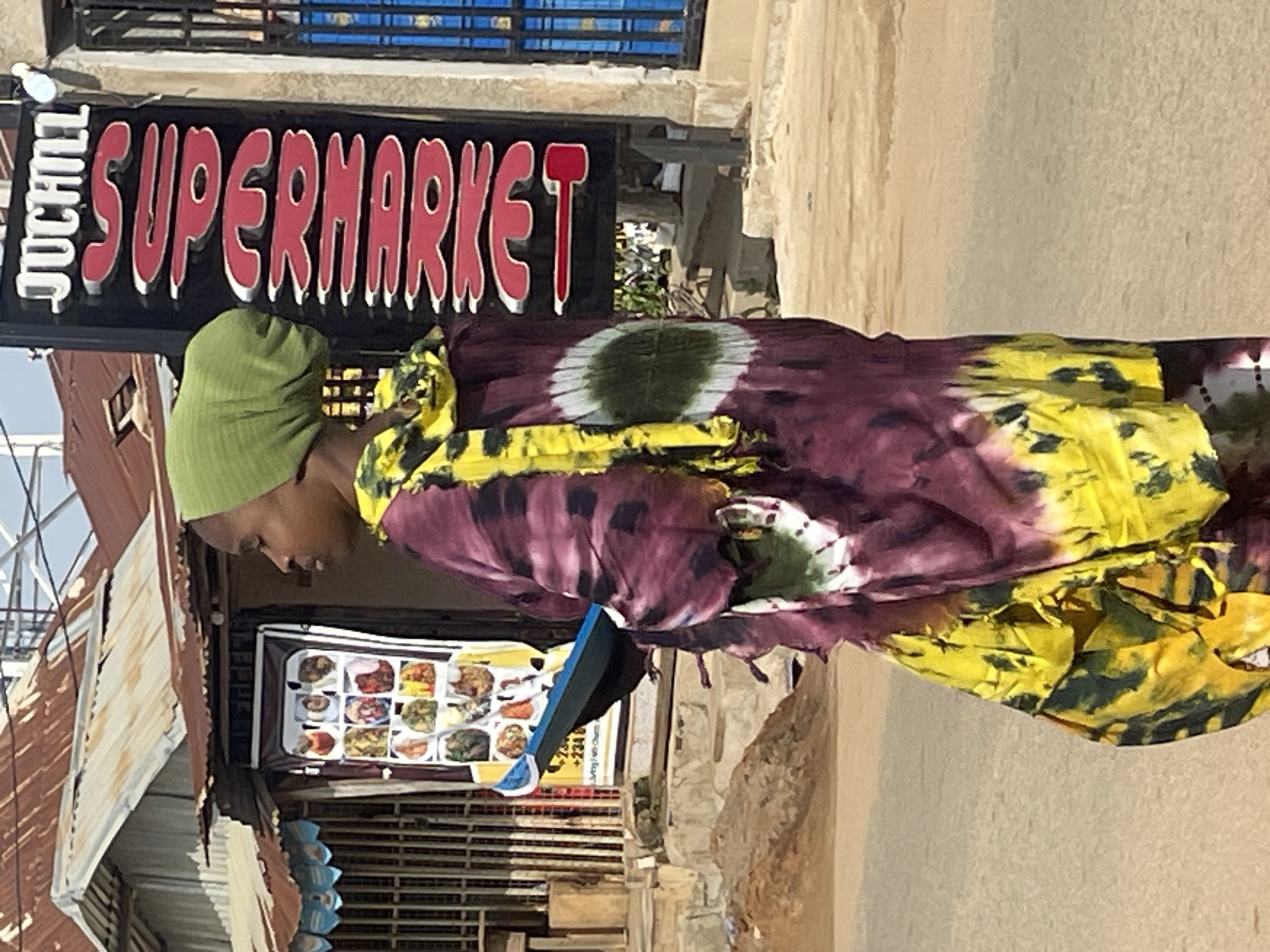 The image depicts a scene in Abuja, Nigeria, focusing on a young woman wearing a green headscarf and a vibrantly colored tie-dye robe. She is positioned in front of a building with a corrugated metal roof, indicative of local architecture. To her right, a vertical sign with red lettering against a black background reads "JUCHILL SUPERMARKET". The woman is holding a dark blue object in her left hand, partially obscured by her attire. Behind her, a display board showcases an array of food items, suggesting the presence of a food vendor or establishment. The environment appears to be an outdoor, possibly street-side location, with a sandy or dusty ground. The overall impression is that of a bustling urban setting within Abuja.