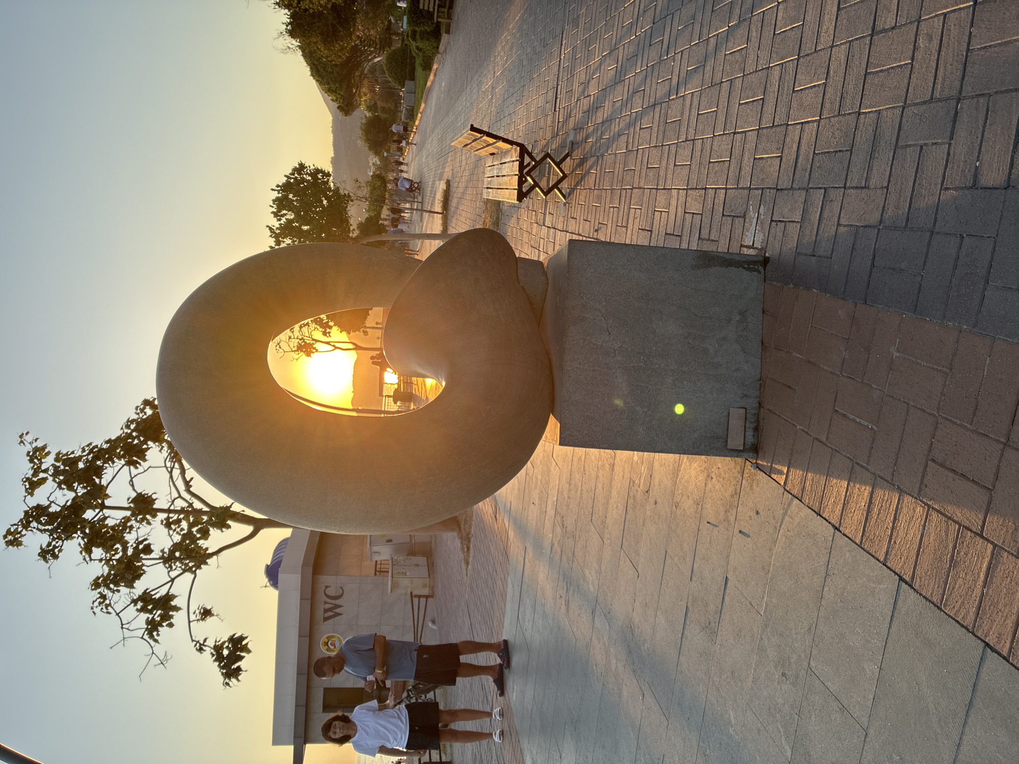 The image captures a sunset scene along a paved walkway in Alanya, Turkey. In the foreground, a large, oval-shaped stone sculpture with a hole through its center is positioned on a stone pedestal. The setting sun shines through the opening of the sculpture, creating a lens flare effect and illuminating the scene with warm, golden light.
To the left of the sculpture, a man and a woman stand near a building with a "WC" sign. The woman wears a white t-shirt and black shorts, while the man is in a grey t-shirt and dark shorts. They appear to be conversing. The building behind them is modern, with tiled walls and a blue dome.
The walkway is paved with interlocking bricks, and in the middle ground, a wooden bench with a dark metal frame sits empty. Further down the walkway, several people are visible walking and strolling, suggesting a public recreational area. Trees with sparse foliage line the right side of the walkway, casting long shadows across the ground due to the low angle of the sun. In the background, a gentle slope with vegetation and distant buildings suggests a hilly terrain. The sky is clear and transitions from a pale orange near the horizon to a light blue above. The overall atmosphere is serene and picturesque, enhanced by the warm glow of the sunset.