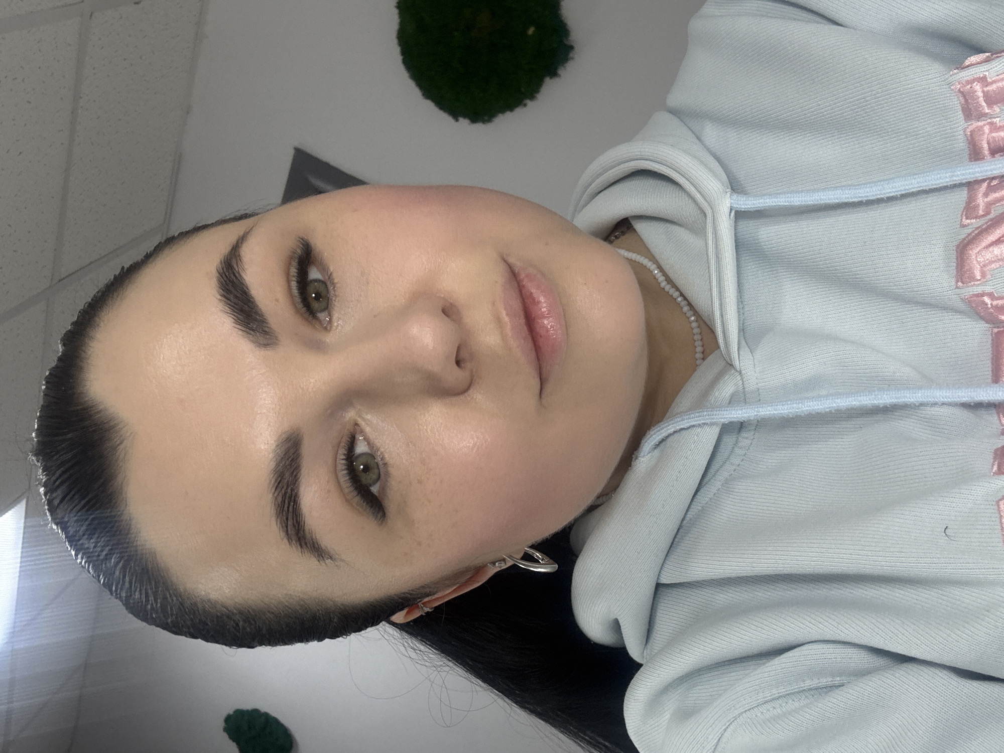 A close-up, angled portrait of a young woman with fair skin, dark eyebrows, and green eyes. She has full lips and her dark hair is styled back. She is wearing a light blue hoodie with pink lettering. A small white beaded necklace is visible around her neck, and she has a silver earring in her left ear. The background is white, with a dark gray trapezoidal shape visible behind her head, and a green moss ball hanging from the ceiling. It is not possible to determine the location in the city of Chaikovsky, Russia, based on this image.
