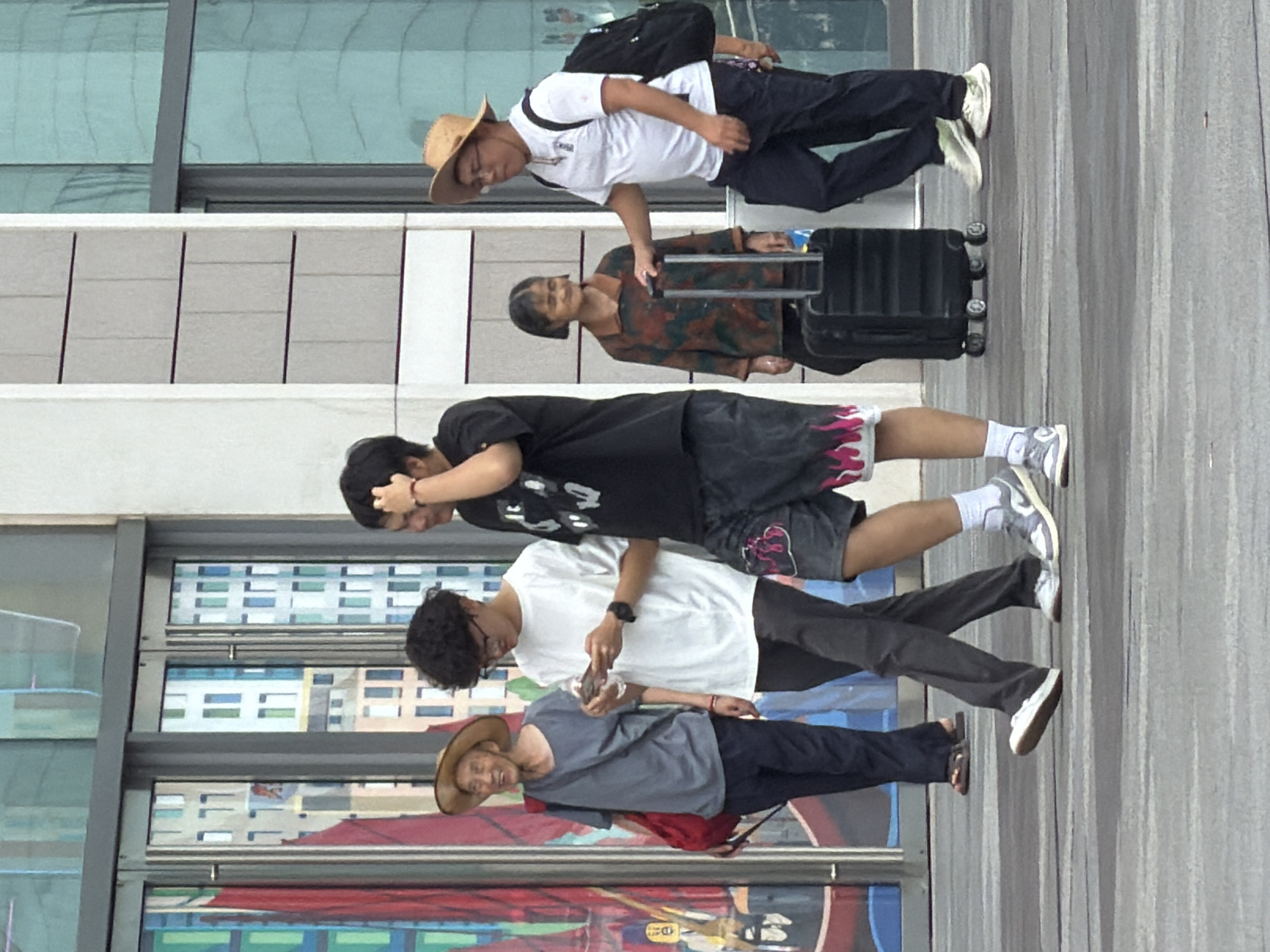 The image depicts a group of people walking on a pedestrian walkway in front of a modern building with large glass windows. The location is identified as the Yuzhong District in China. In the foreground, two young men are walking together. The man on the left wears a white t-shirt and dark pants, and is looking at his phone. The man on the right wears a black t-shirt with graphics and patterned shorts, and has his hand on his head. Behind them, an elderly man wearing a straw hat, a grey t-shirt, and a red bag slung over his shoulder, walks to the left. To the right of the young men, an older woman in a patterned jacket is seen next to a black rolling suitcase, and a young man in a white t-shirt and a straw hat, wearing a backpack, is walking with his hand on the suitcase handle. The environment suggests an urban setting with a focus on the interaction between the individuals and their movement through the space.