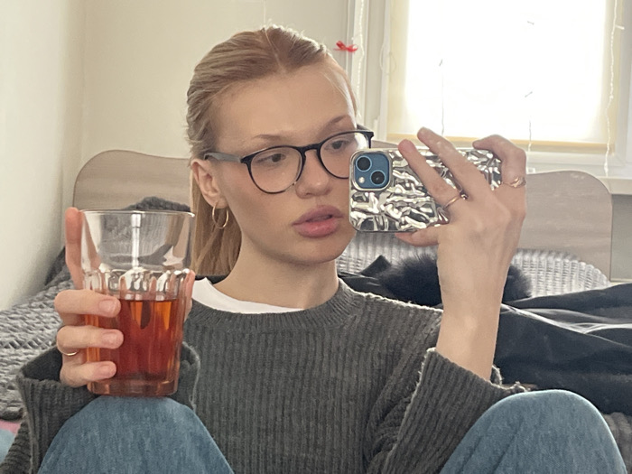 A young female, with fair complexion, light hair pulled back, and black-rimmed glasses, is depicted in an indoor setting in the city of Chaikovsky, Russia. She is wearing a gray ribbed long-sleeve top over a white undershirt and blue jeans. Small gold hoop earrings are visible on both ears. Her left hand holds a clear glass tumbler containing a reddish-brown liquid, likely tea or juice. Her right hand holds a smartphone, specifically an iPhone model with a dual-camera system, encased in a metallic-silver, textured case, positioned as if taking a mirror selfie. She has rings on her left index finger and right ring finger. The background reveals a light-colored wall, a light-toned wooden headboard, a gray textured blanket, and a white object resembling a wicker-patterned pillow. A window with white framing and ambient light is visible in the upper right, and a small red hair accessory is on the upper left wall.