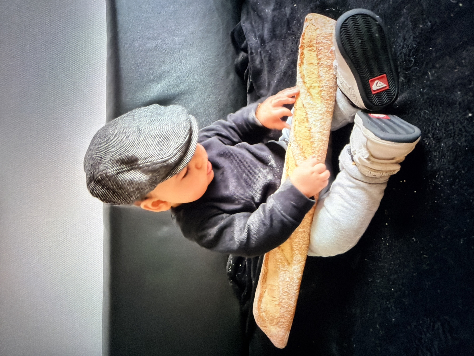 An overhead shot captures a young child, likely a toddler, holding a long baguette with both hands. The child is wearing a grey newsboy-style cap, a dark grey long-sleeved top, and light grey pants. They are also wearing light-colored sneakers with black soles, one of which prominently displays a red Quiksilver logo.
The child is positioned on a dark, textured surface, possibly a couch or a blanket on the floor. Their head is turned to the right, looking down towards the baguette, suggesting curiosity or playful engagement with the bread. The lighting indicates an indoor setting. To the left of the child, a lighter, textured surface, possibly a wall or another piece of furniture, is visible. The overall impression is a candid moment of a child interacting with an oversized baguette.