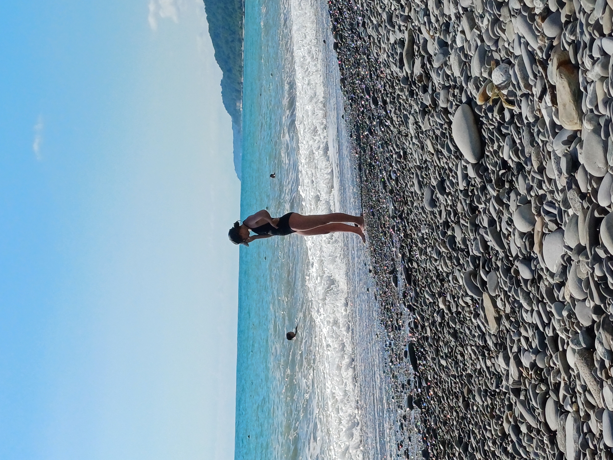 A full vertical shot captures a pebble beach in Sochi, Russia. The beach is covered in smooth, grey stones of varying sizes. In the shallow, turquoise water, a person is partially submerged. Closer to shore, a woman stands facing the water, wearing a black swimsuit and a dark cap. She is looking down. The waves are gently breaking onto the shore. In the distance, a tree-covered mountainside forms the coastline. The sky above is a clear, pale blue with a few wispy clouds.