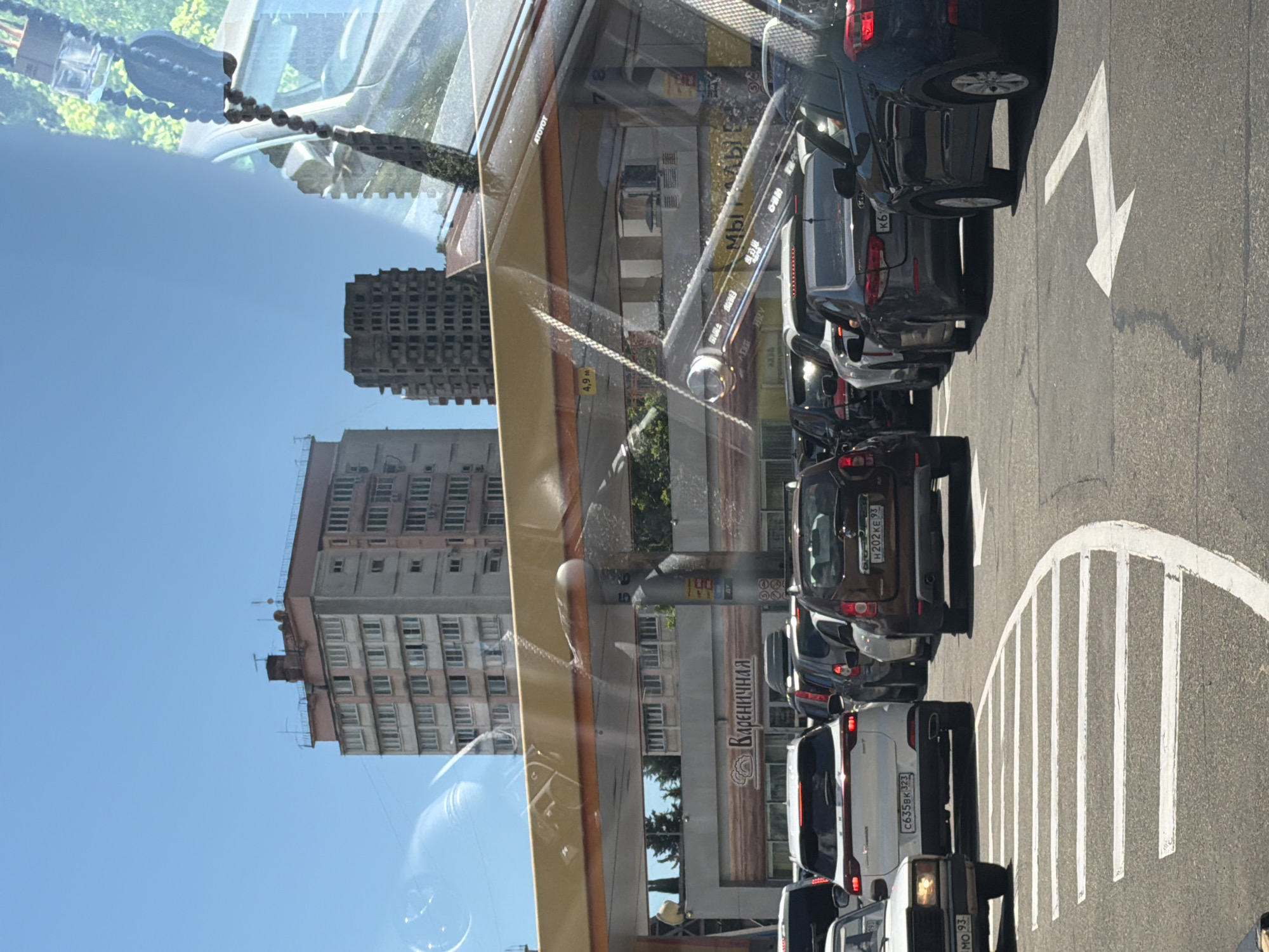The image captures a traffic scene in Sochi, Russia, likely on a sunny day given the clear blue sky. A line of cars is shown, with several vehicles in the foreground and background. The cars are various colors, including black, white, and brown. Some vehicles have visible license plates, such as "H 202 KE 93" and "C635BK 323".
In the background, tall apartment buildings with numerous windows are visible. One building has a sign with Russian text that appears to read "Вареничная". Road markings, including a white arrow indicating a turn and a series of parallel white lines, are present on the asphalt. The scene is reflected in what appears to be a car window, creating a layered effect with greenery and trees visible in some of the reflections. A string of dark, rounded objects, possibly beads or a decorative element, hangs in the upper left portion of the image, also a reflection. The overall impression is of a typical urban environment with everyday traffic.