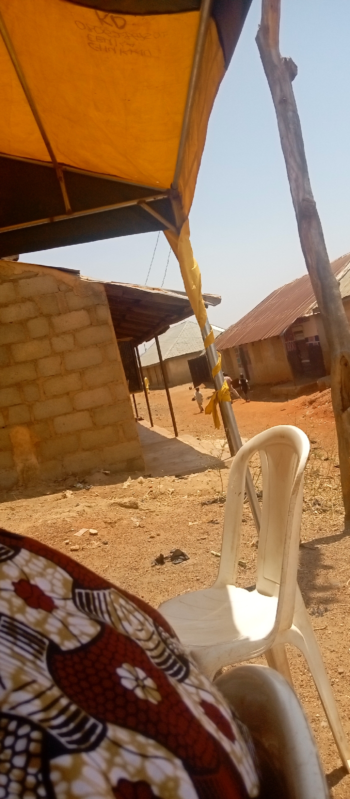 The scene is an outdoor, sunlit area in Kasuwan Magani, Nigeria, characterized by dry, reddish-brown earth. In the immediate foreground, the lower right quadrant reveals a person's lap covered by a patterned fabric featuring red, white, brown, and black motifs. To the right of this, a white plastic chair stands on the dirt. Overhead, a yellow fabric canopy, likely part of a tent structure, is visible, supported by metal poles. Faint text, including "KD" and "ORDER YOURS", is discernible on the canopy's underside.
In the midground, a structure constructed from light-colored concrete blocks with a corrugated metal roof supported by wooden posts stands to the left. A metal pole, partially wrapped in yellow fabric, is prominent, alongside a rough wooden pole. The ground is consistently dusty and dry, with minimal vegetation and scattered debris.
Further in the midground and background, several figures, appearing to be children, are observed on the dirt path between buildings. Multiple single-story residential or commercial structures with corrugated metal roofs, varying in color from silver-grey to rusty brown, line the distant perimeter. The sky above is clear and light blue, indicating bright daylight.