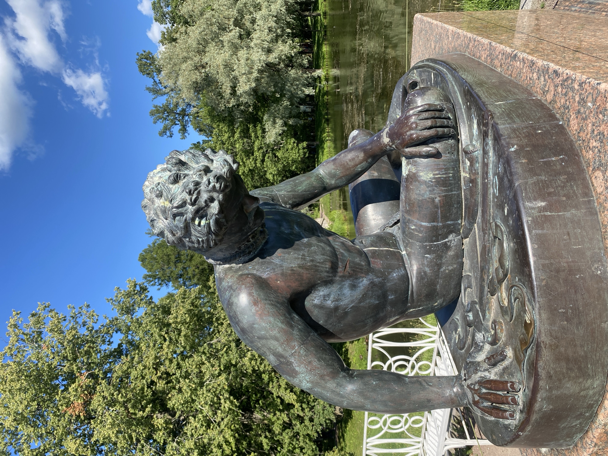 The image shows a bronze statue of a male figure in a park setting. The statue depicts a man with tousled hair, wearing a loincloth, and seated with his head bowed. His body is muscular, and the bronze has a verdigris patina. The statue is positioned on a granite pedestal.
In the background, there is a body of water with lily pads, and beyond that, a lush green landscape with various trees and foliage. A white ornate railing runs along the edge of the water, and a white bench is visible behind it. The sky is bright blue with scattered white clouds.
This statue is part of the Catherine Park in Pushkin, Russia, specifically the "Boy with a Fish" sculpture located near the Grand Cascade. The sculpture is a popular landmark within the park.