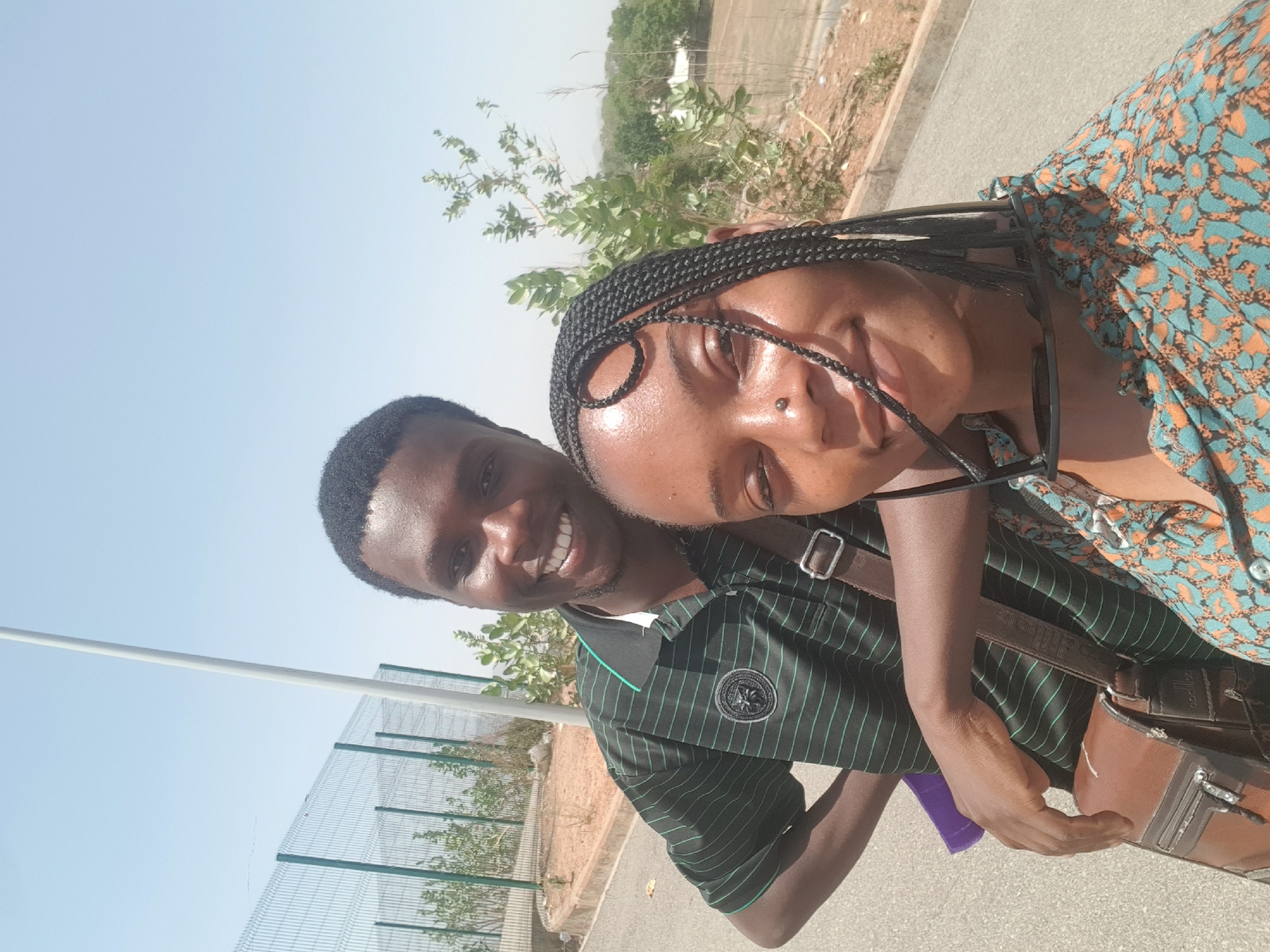 Two individuals, a man and a woman, are posing for a selfie outdoors. The man has short black hair and is wearing a dark green striped polo shirt. The woman has braided hair and is wearing a colorful, patterned top. She also has sunglasses hanging from her neckline. A brown leather bag is slung across their bodies. The background includes a chain-link fence, some trees, and a paved area. It is daytime, and the sky is clear. The location appears to be a roadside or a public area, possibly in Bauchi, Nigeria.