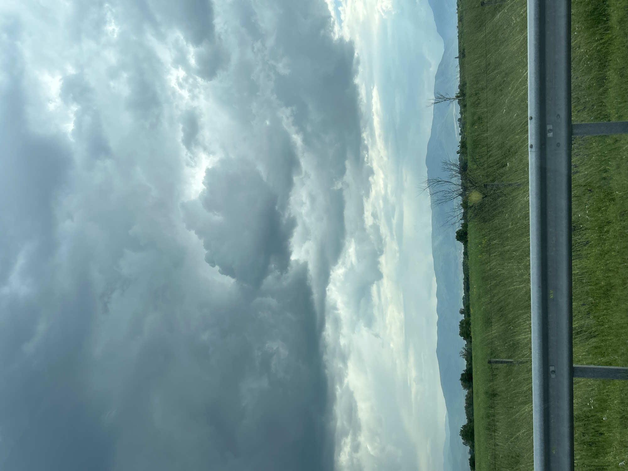 The image shows a landscape scene. The sky is filled with dramatic, dark, and turbulent storm clouds on the left side of the frame, contrasting with brighter, lighter clouds on the right. Below the sky, a green field stretches towards a blurred mountain range in the distance. A gray metal guardrail is visible on the right side of the image, partially obscuring the field. The image appears to have been taken from a moving vehicle. There is no evidence in the image to confirm that this image was taken in Shtarkovo, Bulgaria. There are no visible people, specific actions, or interactions captured in the image.