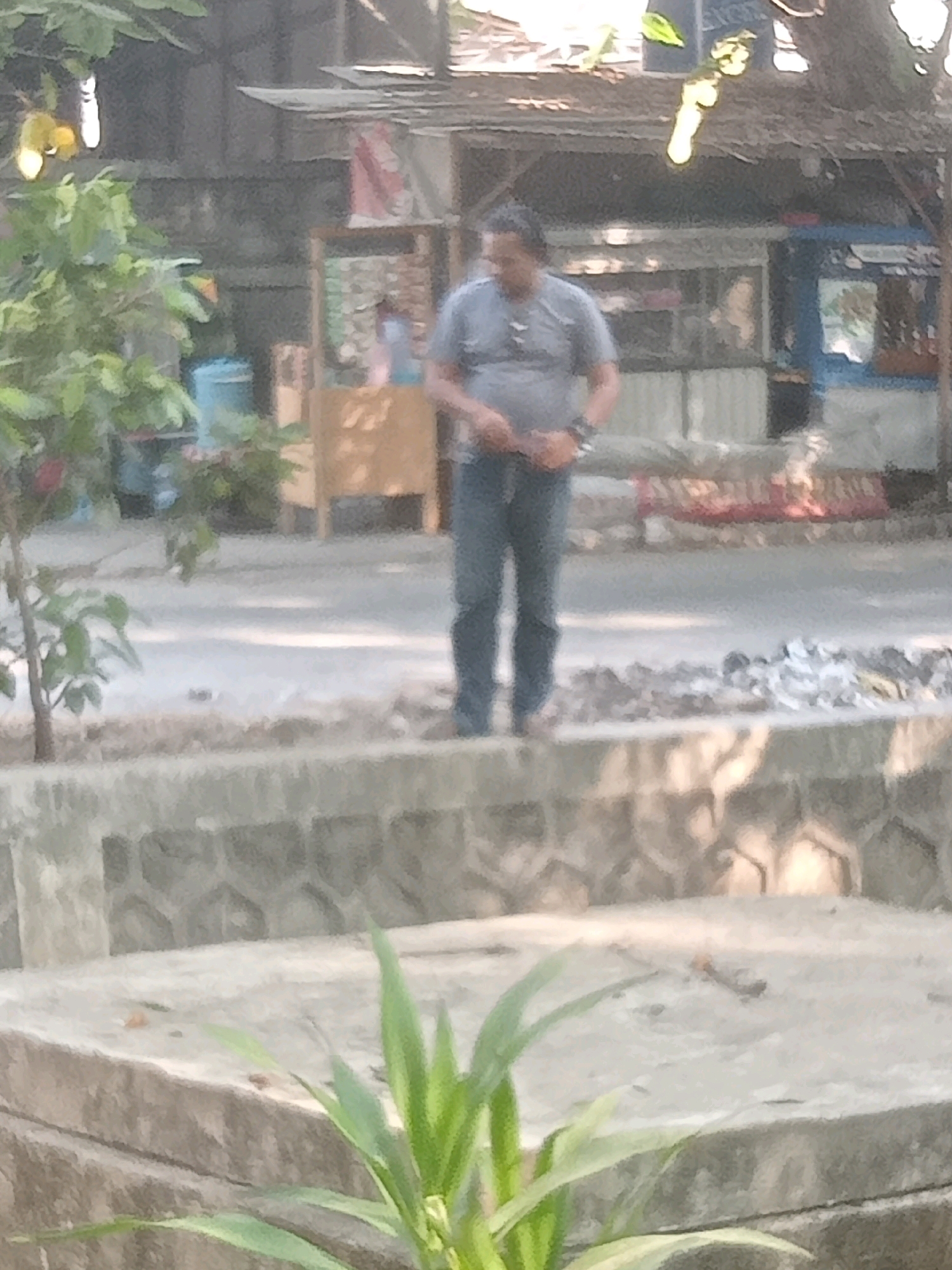 The image captures a scene outdoors, likely in a public area within North Jakarta, Indonesia, given the context. A man, appearing to be of middle age with dark hair, stands in the center of the frame. He is wearing a grey t-shirt and blue jeans. His stance is casual, with his hands clasped in front of him. Behind him, a weathered wooden stall or building with a corrugated metal roof is visible, suggesting a marketplace or roadside establishment. Several other structures and some vegetation are also present in the background, partially obscured by a low stone wall in the foreground. The overall lighting suggests it is daytime.
