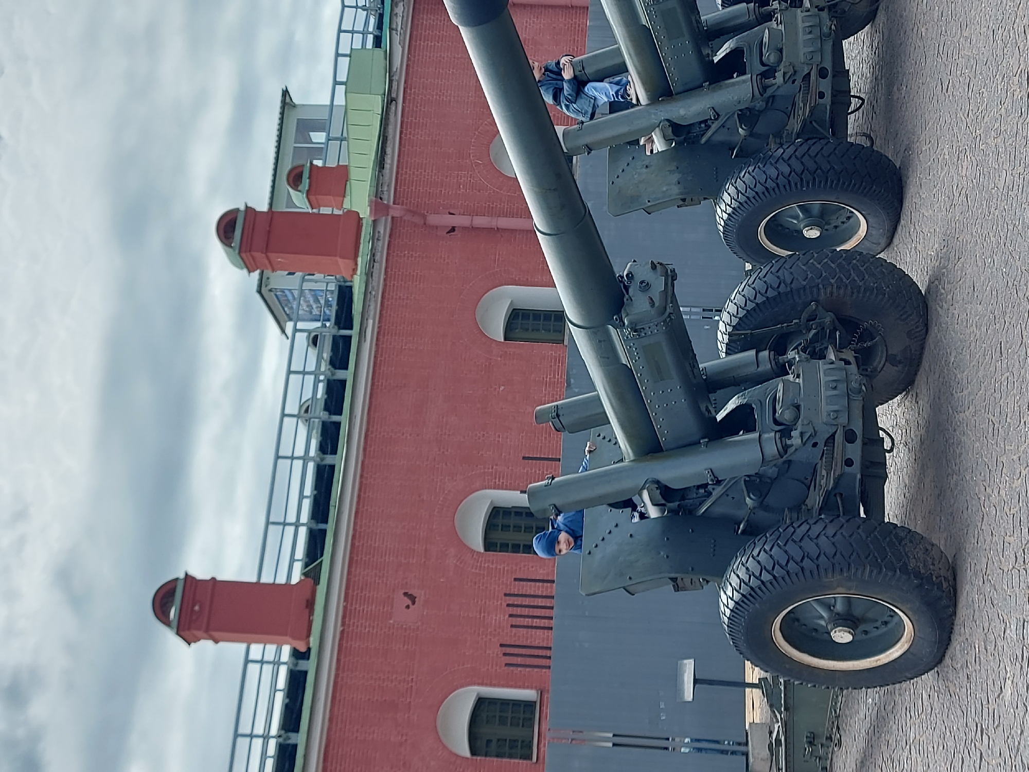 The image shows two cannons displayed in front of a red brick building with white-trimmed arched windows, likely a historical or military museum. A person is sitting on each of the cannons. The sky is overcast. The location is possibly Saint Petersburg, Russia, given the architectural style and the presence of military artifacts.