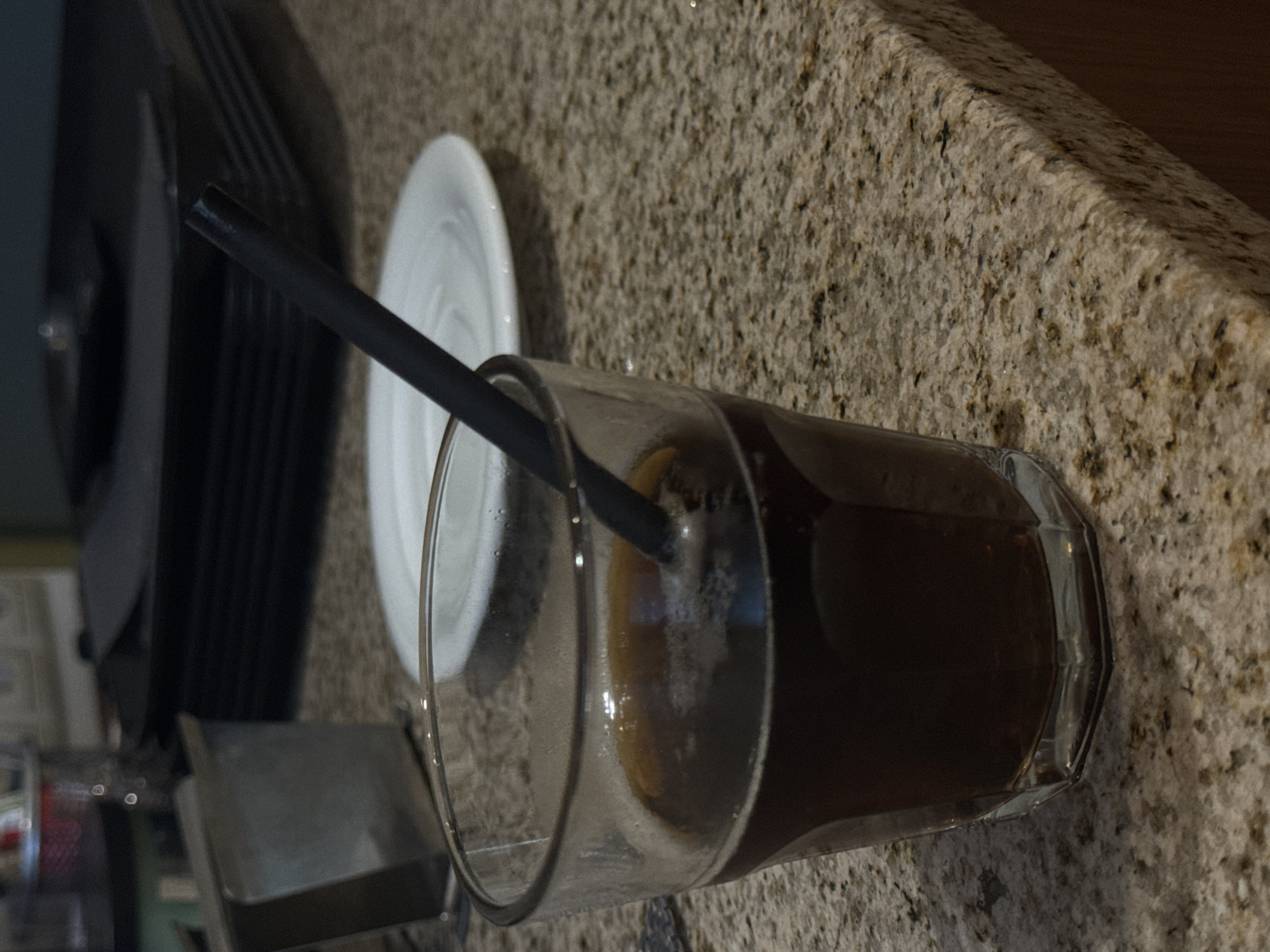 A glass of dark liquid with a straw rests on a speckled granite countertop. In the background, a stack of black rectangular plates is visible, along with a silver napkin holder. The scene is indoors, likely in a kitchen or dining area, with artificial lighting. No specific location in Riga, Latvia can be determined from the image.