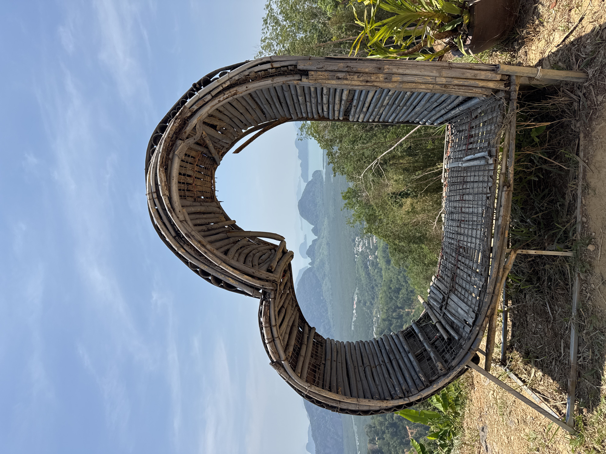 A horizontal, medium-shot photograph shows a large, heart-shaped frame crafted from bamboo and metal against a scenic backdrop. The structure is positioned on an elevated point, framing a landscape of lush green mountains and coastal waters dotted with islands, typical of the southern Thailand region.
The frame itself is constructed from sections of bamboo bound together to form the heart shape. The base of the heart is reinforced with a wire mesh, which likely serves as a platform. The structure is supported by metal bars that act as legs, grounding it on the uneven terrain.
The sky is clear with patches of light clouds. The surroundings are natural with foliage visible to the top and bottom of the image. The photograph is taken during daylight.