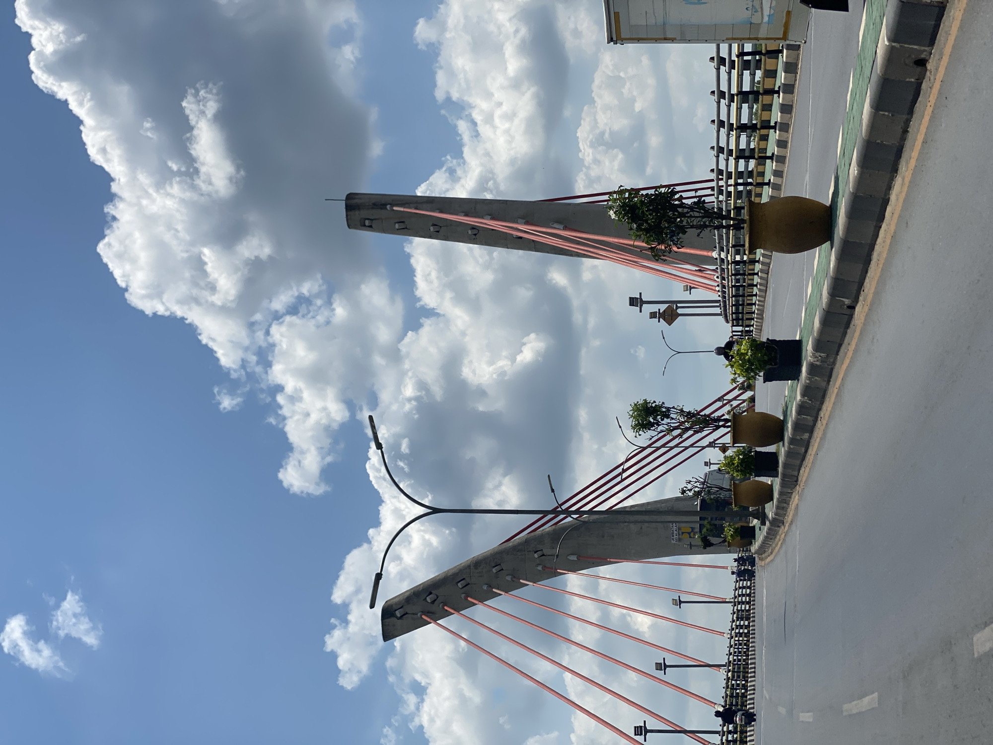 This is a mid-range, eye-level, vertical shot of the Basirih Bridge in Banjarmasin, Indonesia, on a sunny day with scattered clouds. The bridge, a cable-stayed structure, features two distinctive concrete pylons in the background. These pylons are characterized by their angled shape and are adorned with numerous red cables that extend outwards, supporting the bridge. Multiple streetlights with double lamps are visible along the bridge's pathway, and a sign that reads "JEMBATAN DEI ALALAH" is partially visible on the lower section of the right pylon.
The bridge itself has a paved roadway with clear lane markings. Along the edge of the roadway, a decorative green strip is bordered by concrete curbs. Several potted plants, including trees and smaller shrubs, are placed in large, yellowish-brown ceramic pots and black planters, adding a touch of greenery to the scene. A black metal railing with yellow accents separates the roadway from the bridge's pedestrian or safety area.
In the foreground and midground, there are a few people riding motorcycles, indicating activity on the bridge. A white truck is partially visible on the right side of the frame, suggesting ongoing traffic. The sky above is a vibrant blue, dotted with fluffy white clouds, some of which are quite large and dramatic. The overall impression is of a functional urban infrastructure element set against a pleasant, partly cloudy sky.