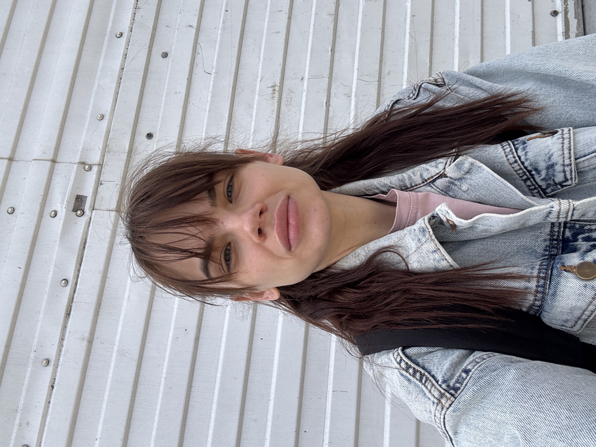 A young woman with brown hair and bangs, wearing a light blue denim jacket over a pink shirt, is posing for a selfie. The background is a white, corrugated metal wall. The scene may be in Rostov-on-Don, Russia.