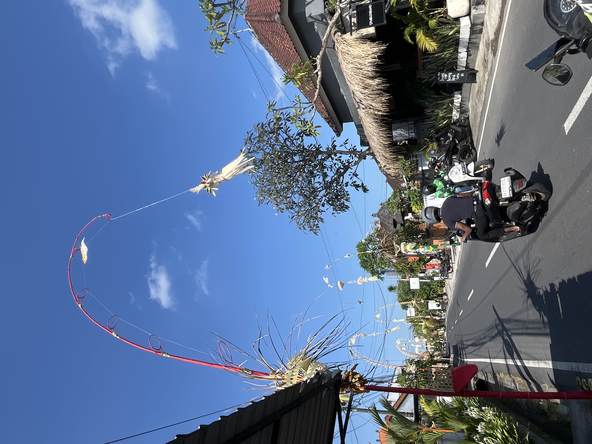 In Mengwi, Indonesia, a street scene unfolds under a clear blue sky with scattered clouds. The road, lined with buildings and vegetation, is occupied by several motorbikes and their riders. One rider, in the foreground, is seen from behind, operating a black motorbike. To their left, another rider in a green jacket is on a white motorbike, and further back, a black motorbike is parked.
The street is adorned with traditional Balinese decorations known as "penjor," which are tall, curved bamboo poles with intricate woven elements and flags, indicating a festive or ceremonial occasion. Several of these penjor arch over the road, with one prominent red and white pole on the left side of the image featuring a woven ornament. Another woven decoration, resembling a bird or kite, hangs suspended from a wire attached to a tall red pole. The buildings visible include a structure with a thatched roof and a tiled roof, and signage suggests commercial establishments. Power lines crisscross the scene above the street and between the buildings. Shadows from the penjor and trees are cast across the asphalt road.