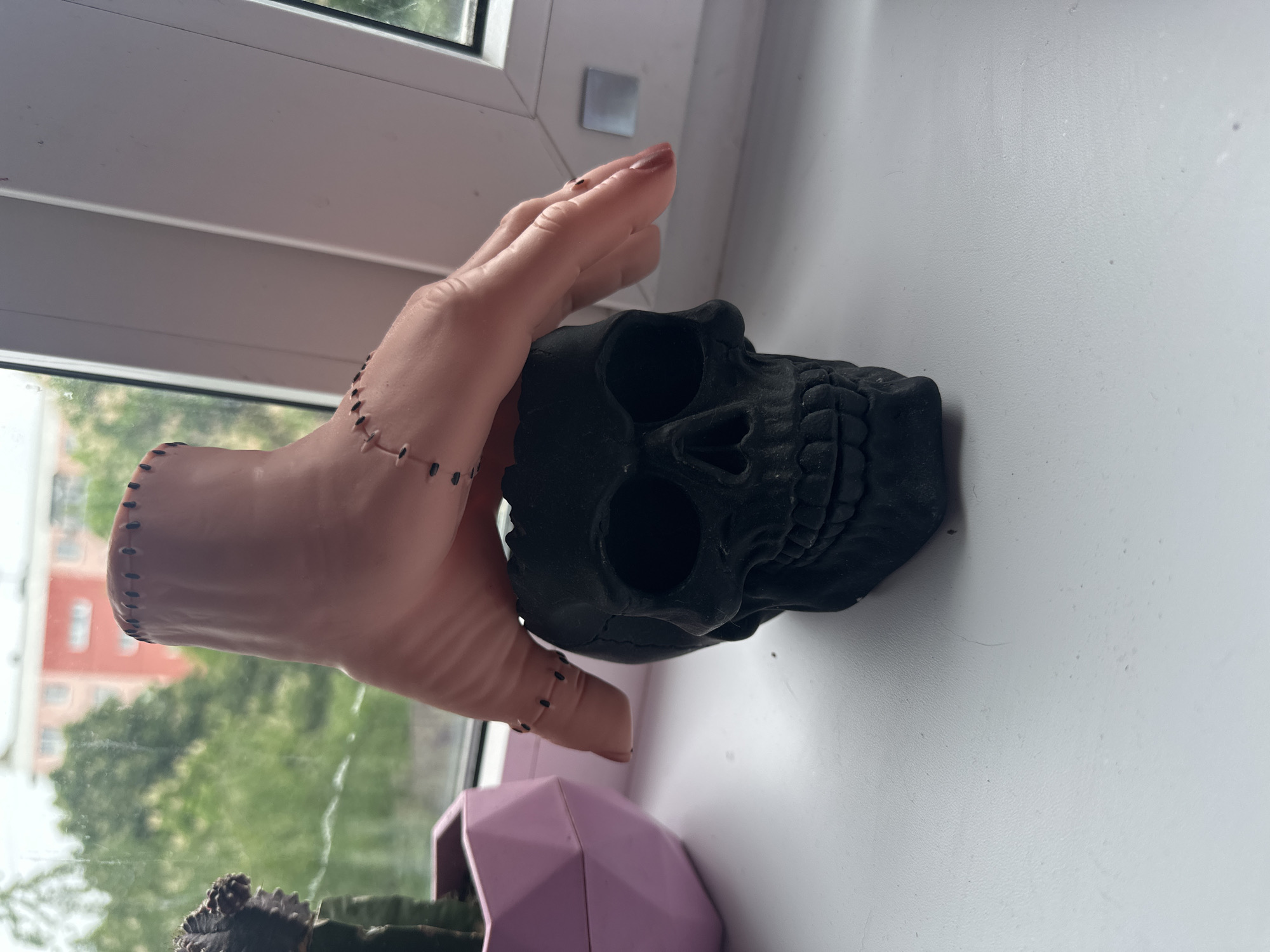 The scene shows a stitched prosthetic hand placed on top of a black skull on a white windowsill. A pink geometric planter with a cactus is visible below the hand and skull. The background shows a blurry outdoor scene with buildings and greenery, suggesting an urban setting. The photograph was taken in Stavropol, Russia.