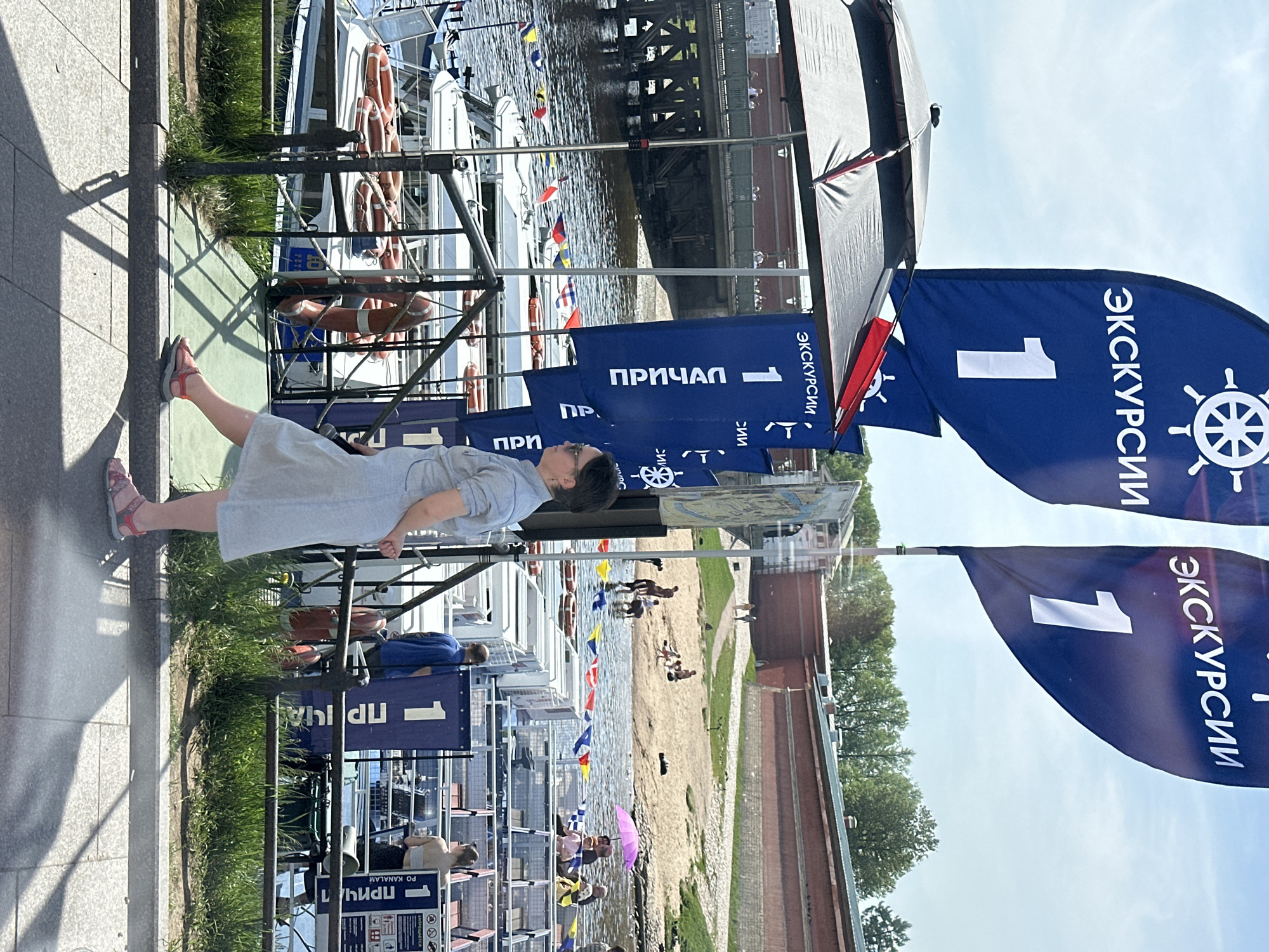 The image shows a woman standing on a gray tiled walkway in St. Petersburg, Russia, with her back to the camera. She is wearing a light blue dress and pink sandals. Behind her, there's a row of boats docked along a riverbank. Each boat is adorned with life preservers. Blue flags with the number "1" and Russian text "ЭКСКУРСИИ" (Excursions) flutter above the boats. The text "ПРИЧАЛ" (Pier) is also visible on smaller signs. A metal fence separates the walkway from the boats. Across the river, there's a sandy beach area with people swimming and relaxing. A brick wall lines the far side of the beach, and trees are visible beyond the wall. A bridge spans the river in the background. The sky is light blue, suggesting it is daytime.