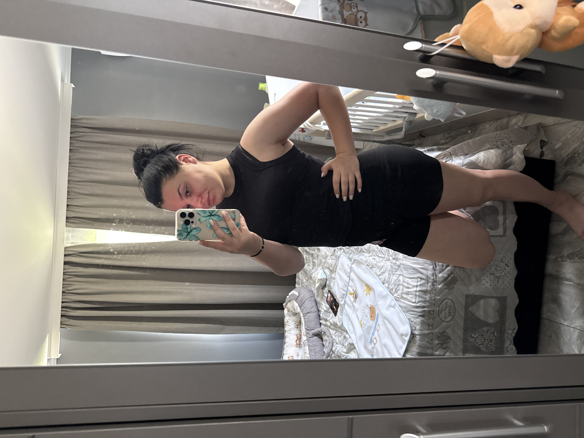 A woman with dark hair pulled up in a bun is taking a mirror selfie in a room. She is wearing a black sleeveless top and black shorts. Her right hand is on her hip, and she is holding a phone in her left hand. In the reflection, a crib and a baby mattress are visible. The room has grey curtains and a grey dresser. There is a stuffed monkey toy hanging from the dresser. The image is taken inside a home, likely in a bedroom or nursery. The location is stated to be in Rogosh, Bulgaria.