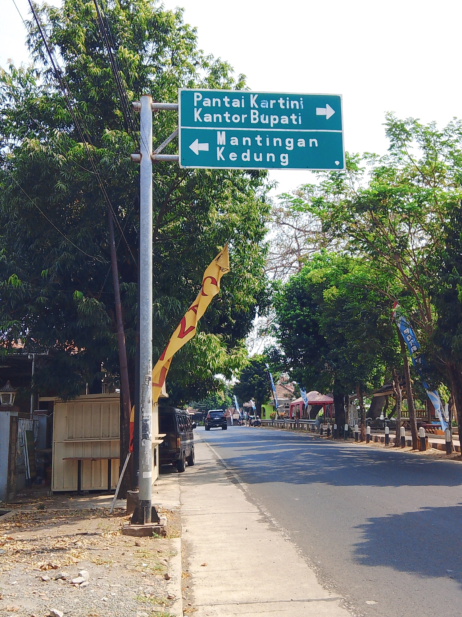 The image shows a street scene in Potroyudan, Indonesia, likely during daytime. A green directional sign is prominently displayed on a metal pole, indicating "Pantai Kartini" and "Kantor Bupati" to the right, and "Mantingan Kedung" to the left. The sign is partially obscured by the pole and trees.

To the left of the sign pole, there is a sidewalk area with scattered dry leaves and some small rocks. Adjacent to the sidewalk is a light-colored, modular structure, possibly a kiosk or vendor stall, with shelves visible inside. A dark-colored van or minibus is parked behind this structure.

The street itself is paved and appears to be a two-way road. Trees line the right side of the street, casting shadows on the asphalt. In the distance, there are buildings, tents or awnings, and other vehicles. The overall environment suggests a semi-urban or suburban setting. A yellow flag with red markings is attached to the sign pole and flaps in the wind.