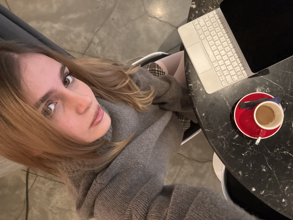 A high-angle photograph captures a young adult female seated at a black marble-patterned round table within an indoor public establishment, likely a cafe or coffee shop, in the city of Unknown, Unknown. The female subject has light brown hair and is wearing a grey, high-neck knitted sweater and a brown plaid skirt, partially obscured by the table. She is looking directly upward into the camera. On the table, to the right of the subject's position, rests a red cup on a matching red saucer containing a light brown beverage, accompanied by a dark packet and a blue stirrer. Adjacent to the cup, a white electronic keyboard with an attached black screen is positioned. A white chair is partially visible behind the subject. The floor visible in the background is light grey concrete with visible crack lines.
