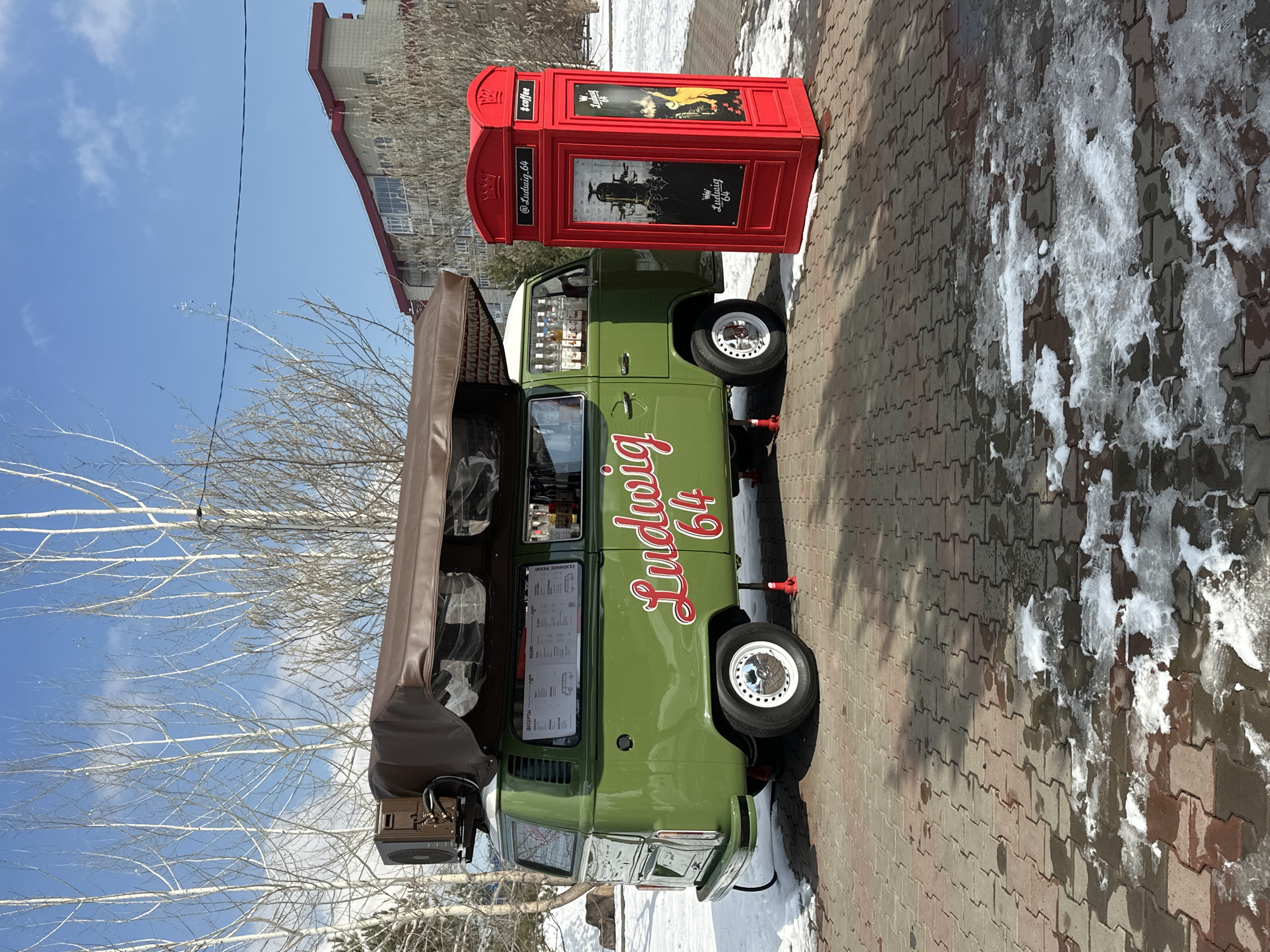 The image shows a green "Ludwig 64" van parked on a brick-paved area next to a red phone booth-shaped kiosk. The van has a brown awning extended, and there is snow on the ground. The background includes a building and a clear blue sky. The location is Krasnoyarsk, Russia.