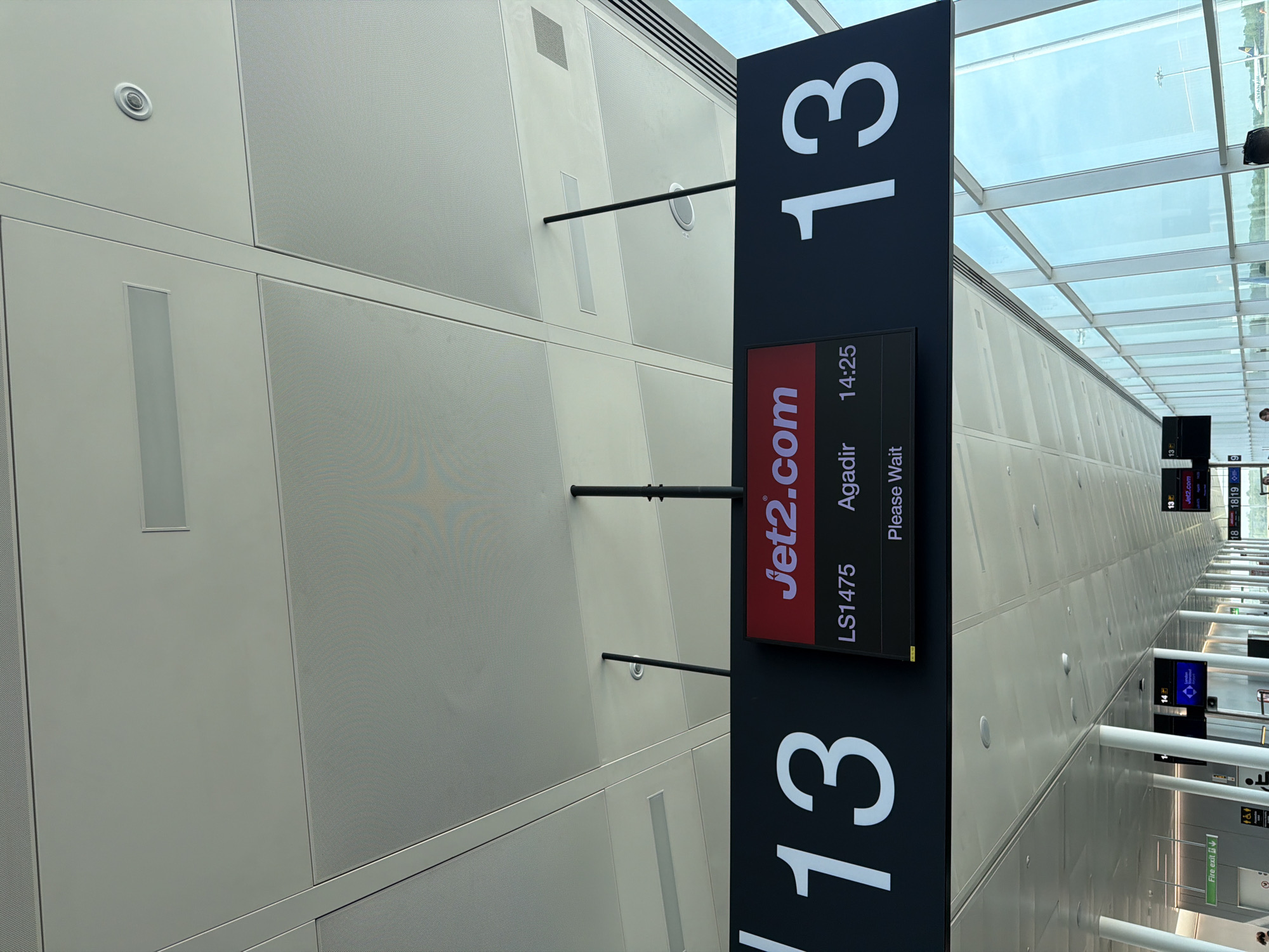 The image shows a sign at an airport in Uttlesford, United Kingdom, indicating gate 13 for a Jet2.com flight LS1475 to Agadir, departing at 14:25. The sign displays "Please Wait". The airport has a modern design with a glass ceiling allowing natural light to enter. Other gate signs can be seen in the distance, along with support columns. The wall is composed of rectangular panels with a perforated texture.