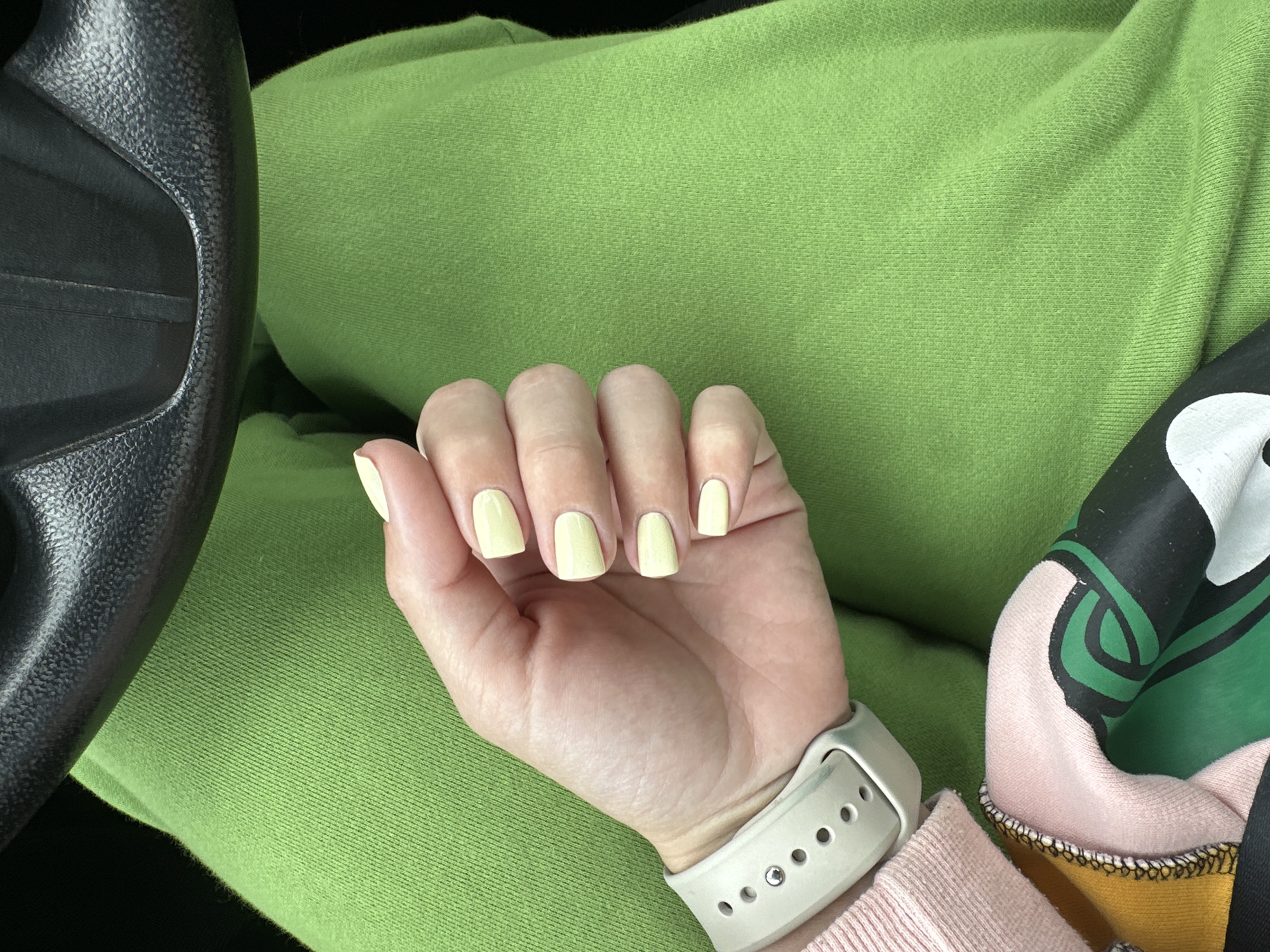 A person's hand with light yellow painted nails rests on their lap, which is covered by bright green fabric. The hand is wearing a light gray watch band. A portion of a black steering wheel is visible in the upper part of the image. The clothing is a light pink sleeve peeking out from beneath the green fabric. The image context does not contain information about the city of Perm, Russia.