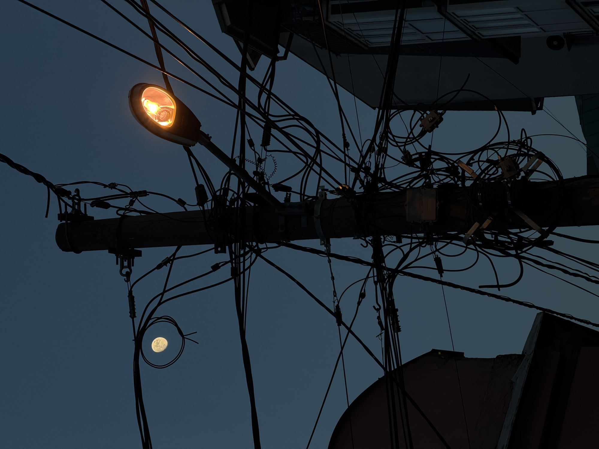 The photograph depicts a cluttered urban scene, likely in Palilula Urban Municipality, Serbia, given the region's characteristic power line configurations. A utility pole is central, supporting a street lamp with an illuminated bulb. A dense network of tangled electrical and communication wires extends from the pole, obscuring the view of the sky. In the background are building structures, possibly residential or commercial, with visible windows and rooftops. In the lower-left quadrant, the moon is visible through a gap in the wires. The overall mood is dusky, suggesting either early evening or twilight.