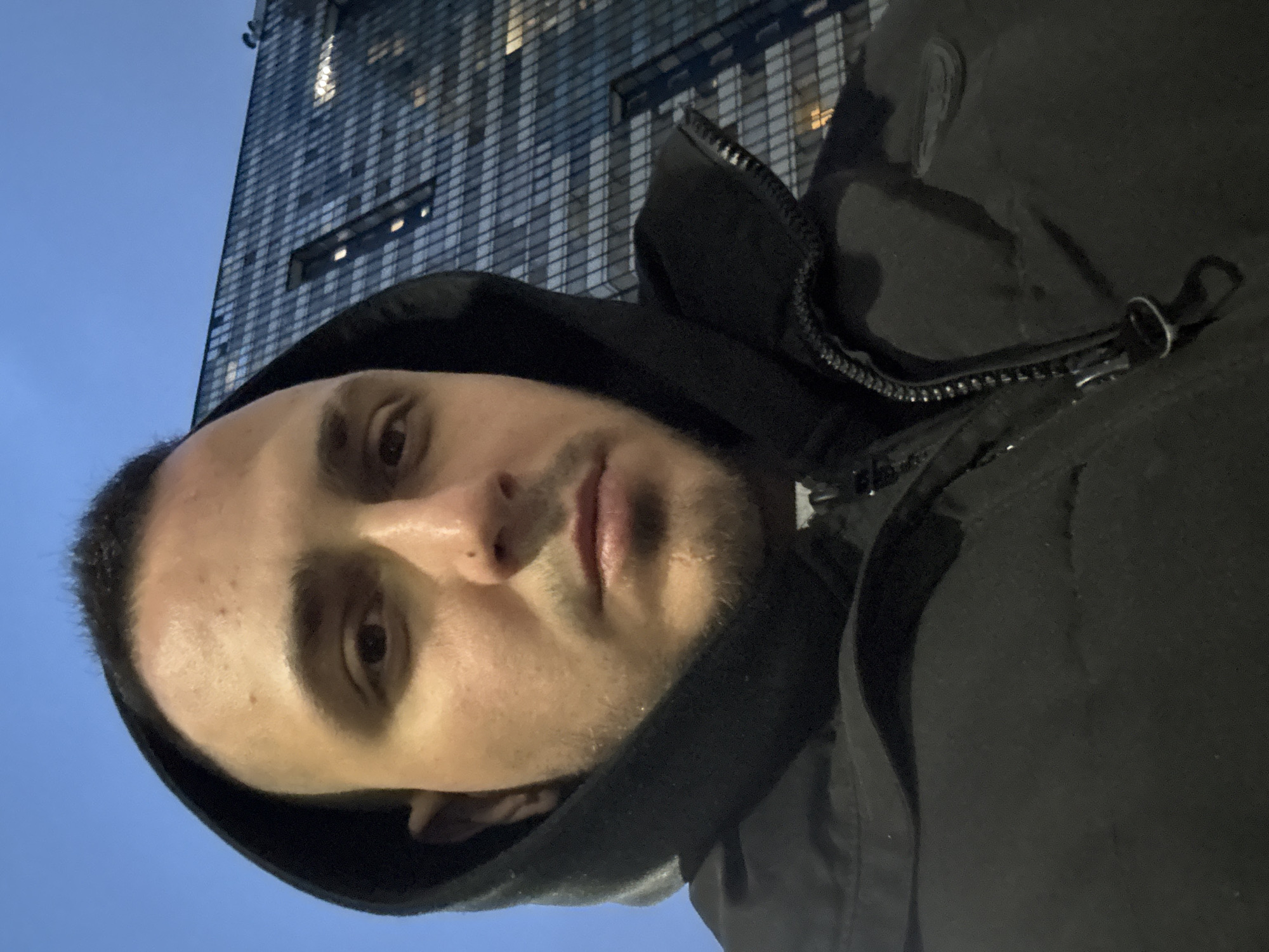 A portrait of a man is presented against the backdrop of a tall building with a gridded exterior, likely at night. The man is wearing a black hooded jacket, with the hood up. The jacket is partially zipped. The building features lighted windows. The sky is a gradient, suggesting dusk or dawn. The location is in a city of Unknown, Unknown.