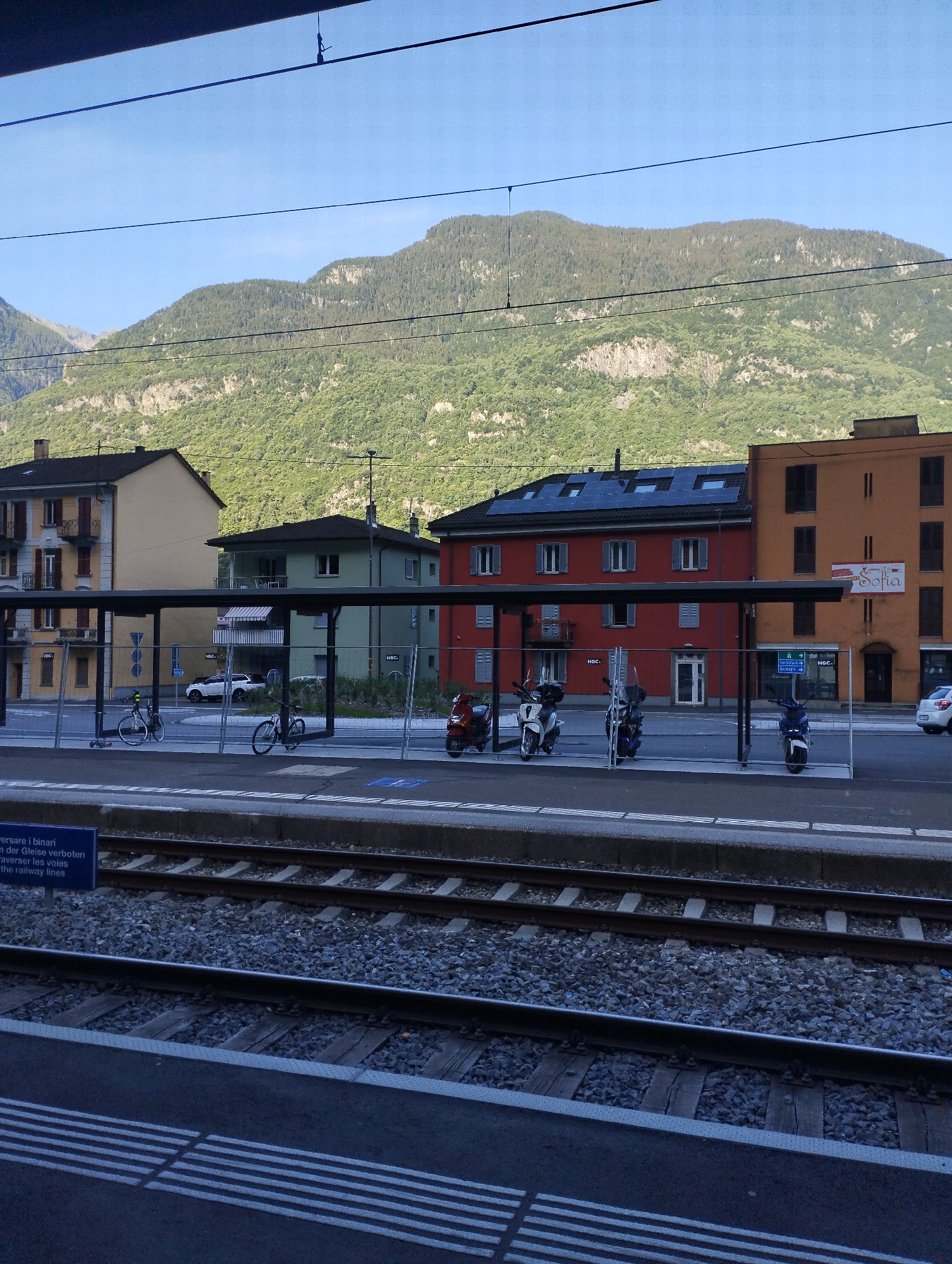 An outdoor, eye-level, medium shot captures a train station platform with a backdrop of buildings and a forested mountain under a clear blue sky. The platform is made of concrete and gravel, with train tracks running parallel to it. On the platform, two bicycles are parked. Several motorcycles are also present, including two white scooters and a blue one. Behind the platform, a row of multi-story buildings can be seen. The building on the left is pale yellow with brown trim, the middle building is red with a dark roof and solar panels, and the building on the right is orange. The mountain, covered in dense green trees, rises steeply behind the buildings. A blue sign with white text is visible in the lower left corner of the image, partially obscuring the train tracks. A white car is parked in front of the orange building. The lighting suggests it is daytime.