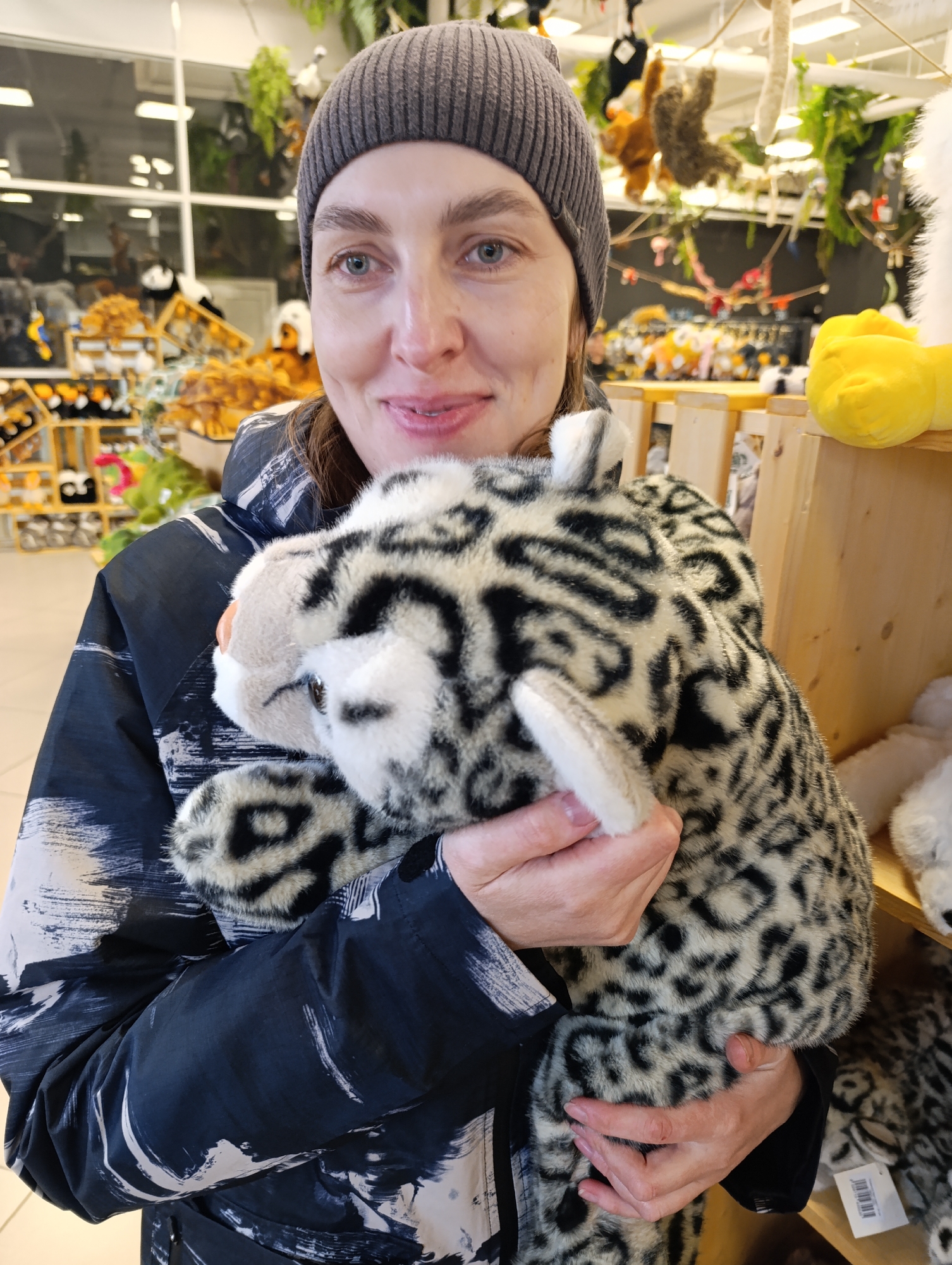 A woman wearing a gray beanie and a black and white jacket is holding a stuffed snow leopard in her arms. The background shows a store setting with shelves displaying various stuffed animals and toys. A window in the background provides natural light, and hanging stuffed animals are visible from the ceiling. The location is likely in Perm, Russia, as indicated in the instructions, but no specific details in the image confirm this.