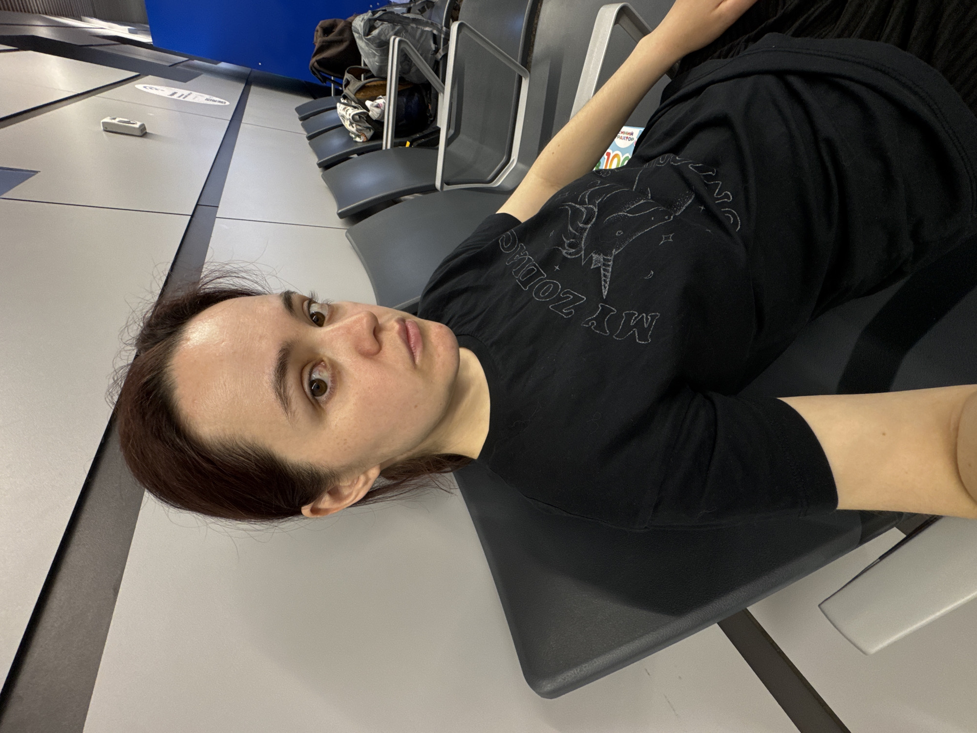 A young woman is lying on airport seating. The woman has light skin and auburn hair. She is wearing a black t-shirt. The seating is gray. Behind her is more airport seating and luggage. A blue ceiling is visible in the background. There is a sign on the wall that says "РОССИЙСКИЕ АВИАЛИНИИ." The scene appears to be at Sochi International Airport in Russia.