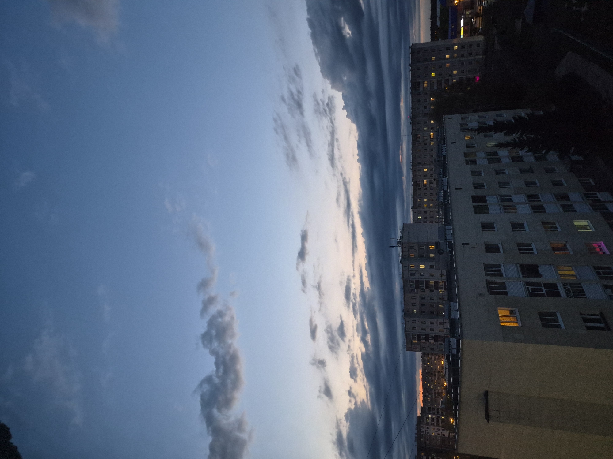The image captures an evening view in Tobolsk, Russia, showcasing residential buildings illuminated with scattered lights against a dusk sky. The buildings are multi-storied and display a repetitive window pattern. Dark, striated clouds dominate the skyline, suggesting a possible storm. The lighting in the buildings indicates inhabited spaces, with a few windows displaying colorful light. The surrounding environment appears urban, with other buildings visible in the distance.