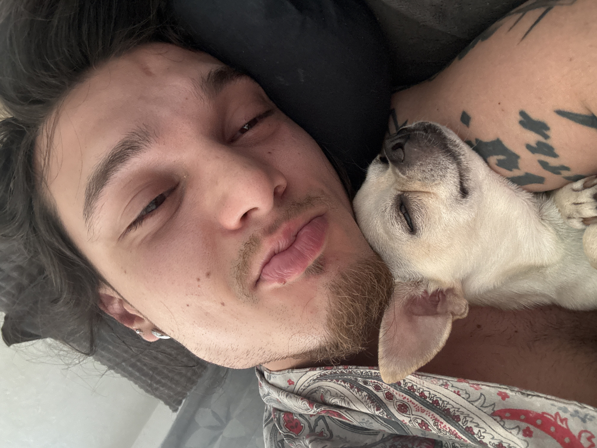 A selfie-style, close-up photograph features a man lying down with a dog nestled against him. The man has fair skin, dark hair and eyebrows, and a goatee. His eyes are partially closed. He is wearing an earring in his left ear, and a patterned garment is visible around his neck and chest. His right arm, also fair-skinned, is visible behind the dog and features dark tattoos.
A small, light-colored dog with closed eyes is positioned so that its head rests on the man's arm and chest.
The location is undetermined, but not necessarily Sofia, Bulgaria. The setting appears to be indoors and the background is partially visible, consisting of neutral and dark colors. The interaction suggests a scene of companionship and rest.