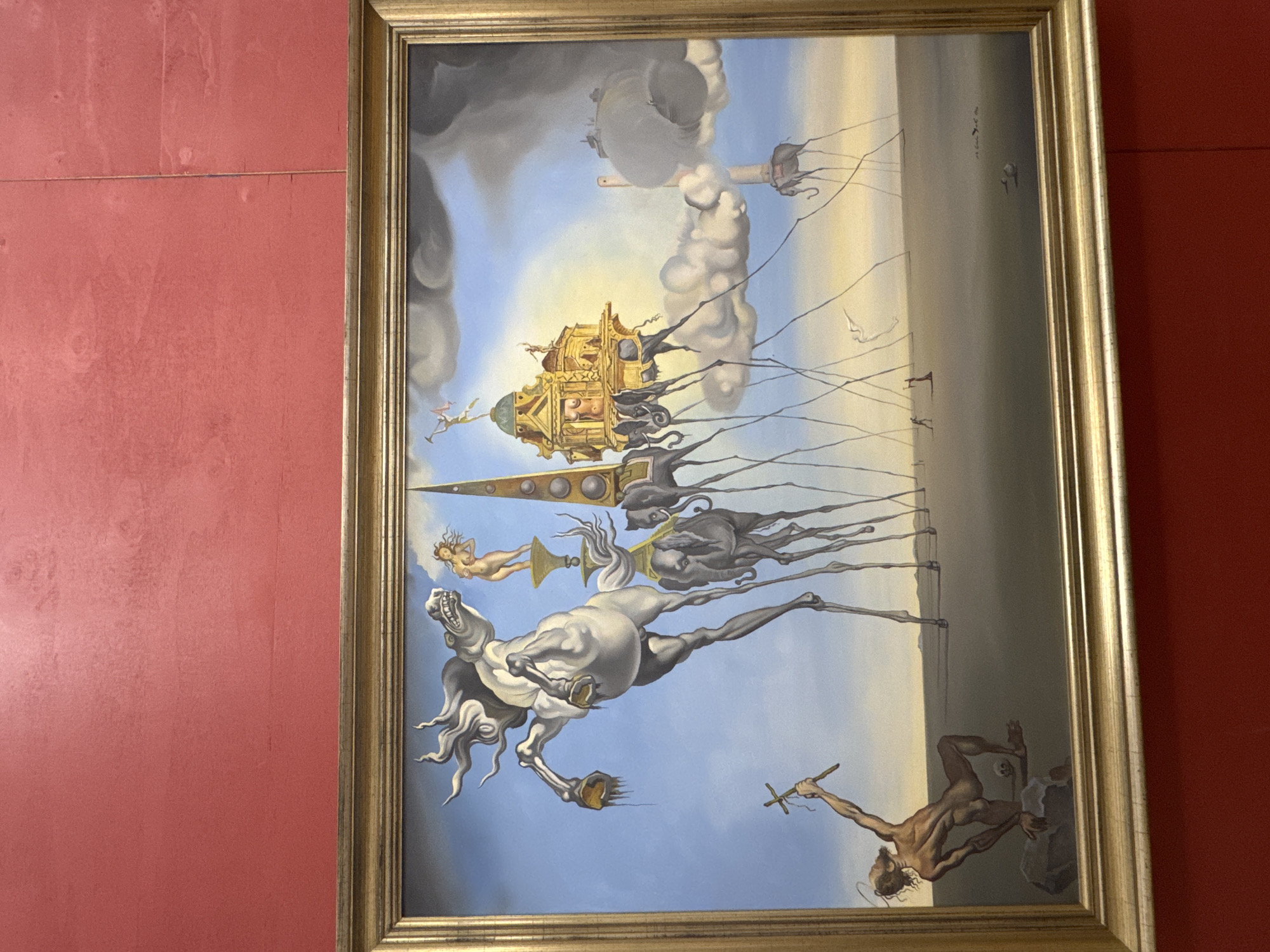 The image displays a framed surrealist painting. The painting depicts a barren, sandy landscape under a blue sky with scattered clouds. In the foreground, a muscular, emaciated figure kneels on a rock, holding a cross aloft. To the left, a white horse rears up on impossibly thin legs, its mouth open in a snarl, with dripping objects from its hooves. In the mid-ground, several elephants with elongated, spindly legs walk across the desert. One elephant supports a gilded, architectural structure resembling a building or temple, topped with a small figure. Another elephant is connected to a cloud by a thin rope. In the far distance, a figure stands silhouetted against the horizon, and other distant structures are visible. The painting is a representation of "The Elephants" by Salvador Dalí. The painting is displayed on a red wall. There is no information in the image to suggest it is located in the city of Pernik, Bulgaria.