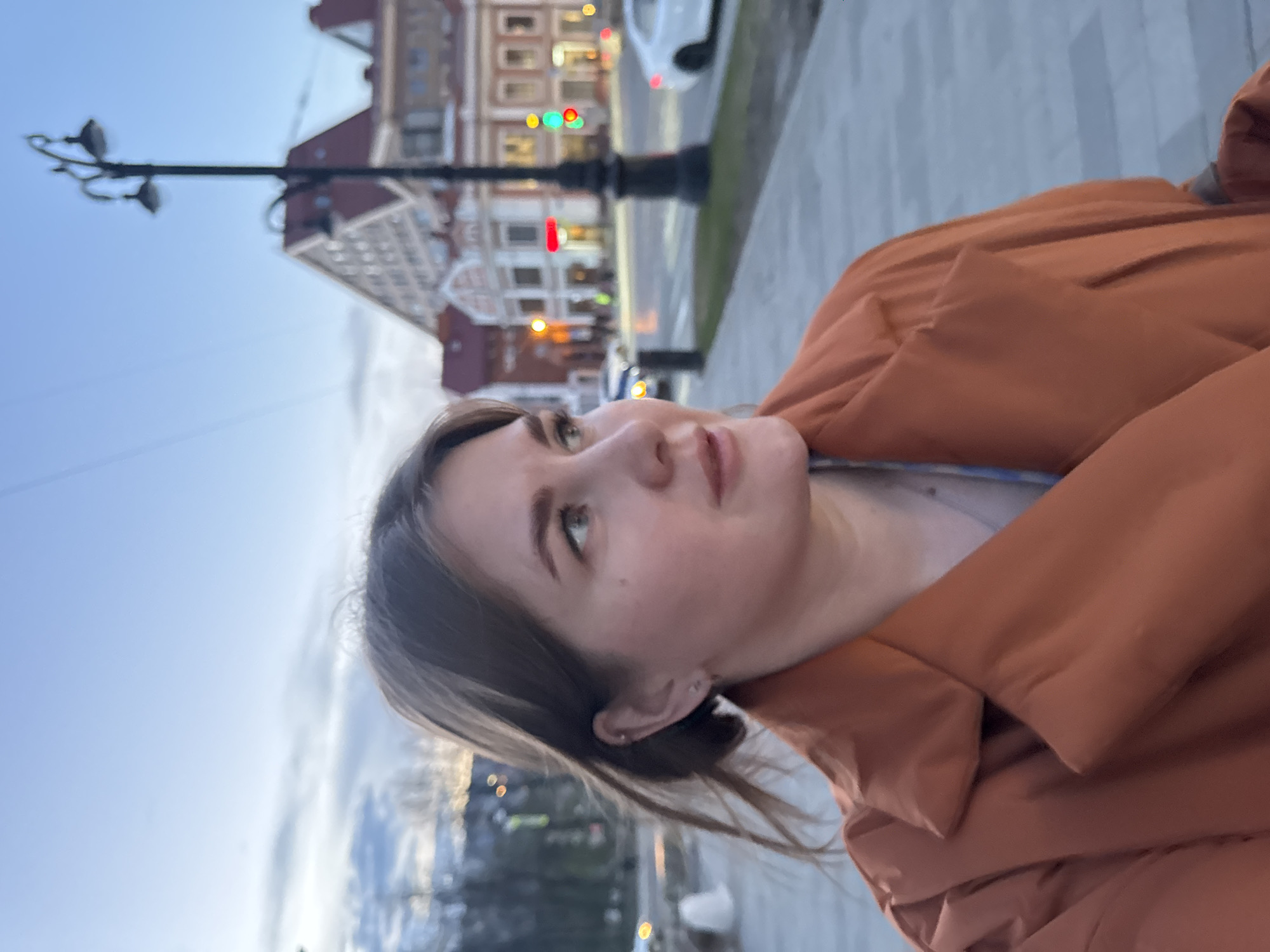A woman wearing a brown jacket is the primary subject of this outdoor photograph. She is positioned on a sidewalk, with a city street in the background. Notable elements in the background include a street lamp and a building in the city of Perm, Russia. The image is rotated counter-clockwise.