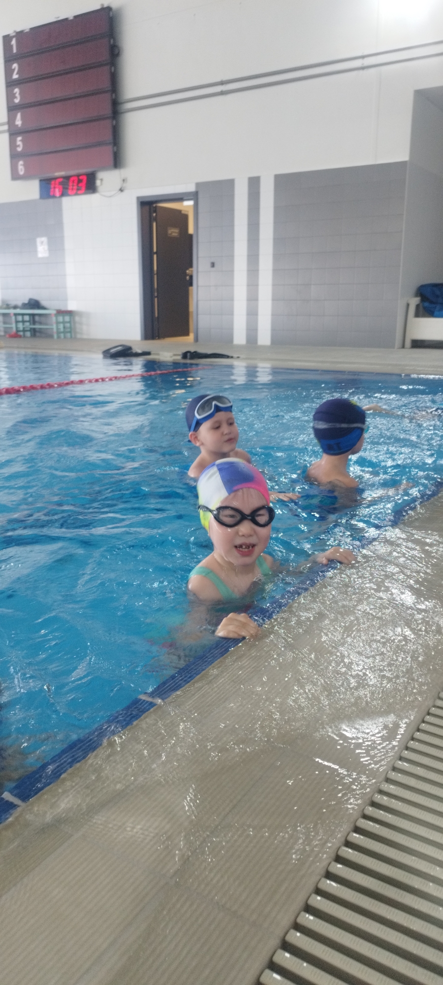 Three children are in a swimming pool. Two are wearing swim caps and goggles. One is holding onto the edge of the pool. The pool has a lane rope. A digital clock on the wall shows 16:03. The location is a swimming pool in what appears to be a gym or public athletic facility. There is a sign with numbers 1-6 on the wall. The exact location in the city of Yakutsk, Russia cannot be determined from the image.